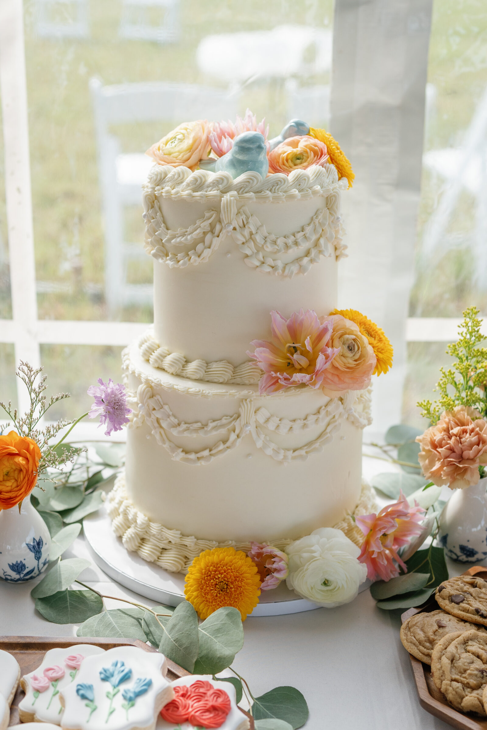 A two-tier wedding cake decorated with piped swags and fresh pastel flowers on a reception dessert table.