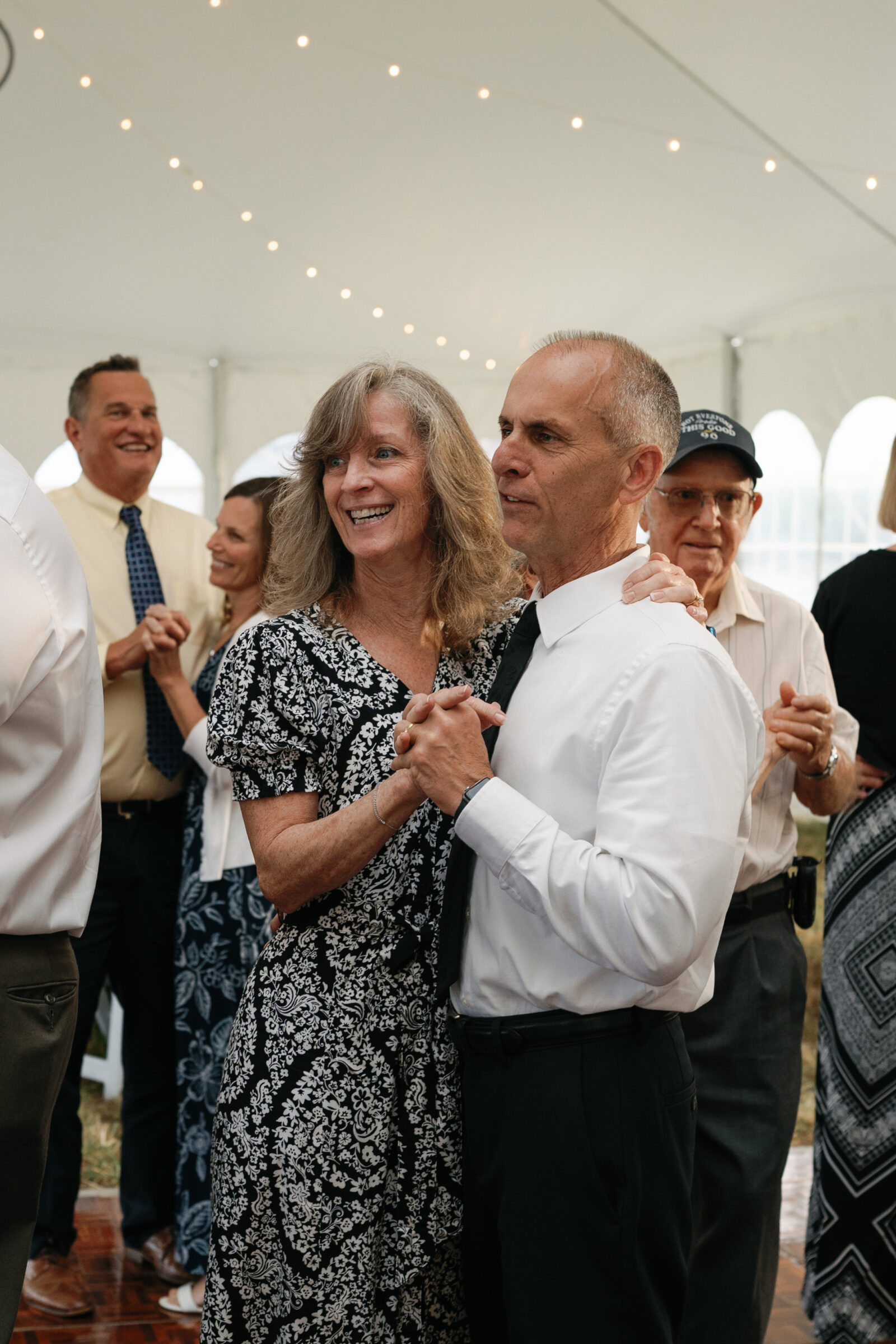 A couple smiles while dancing together during the reception, surrounded by guests under the tent lights.