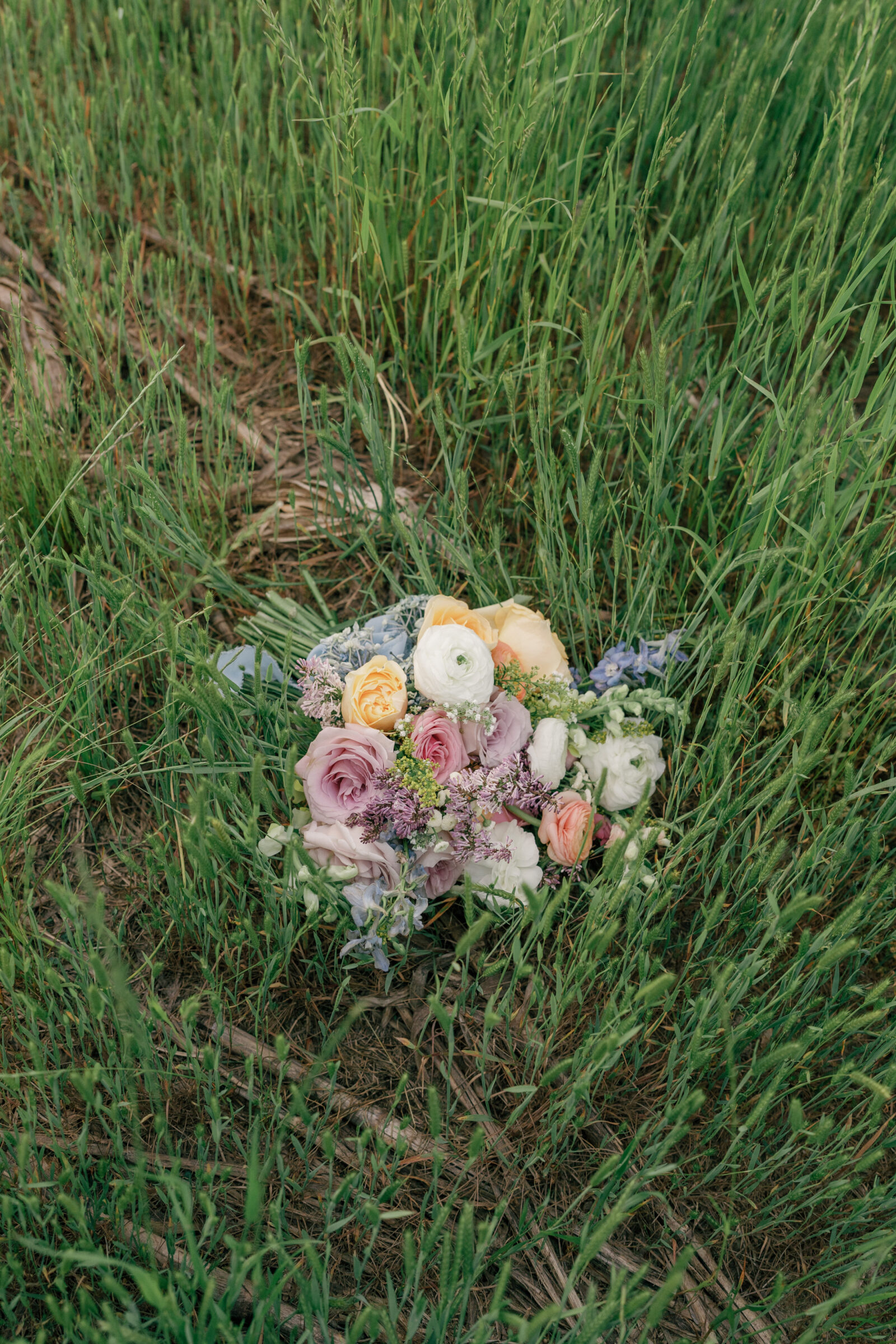 Bridal bouquet with pastel roses, ranunculus, and wildflower accents resting in tall green grass.