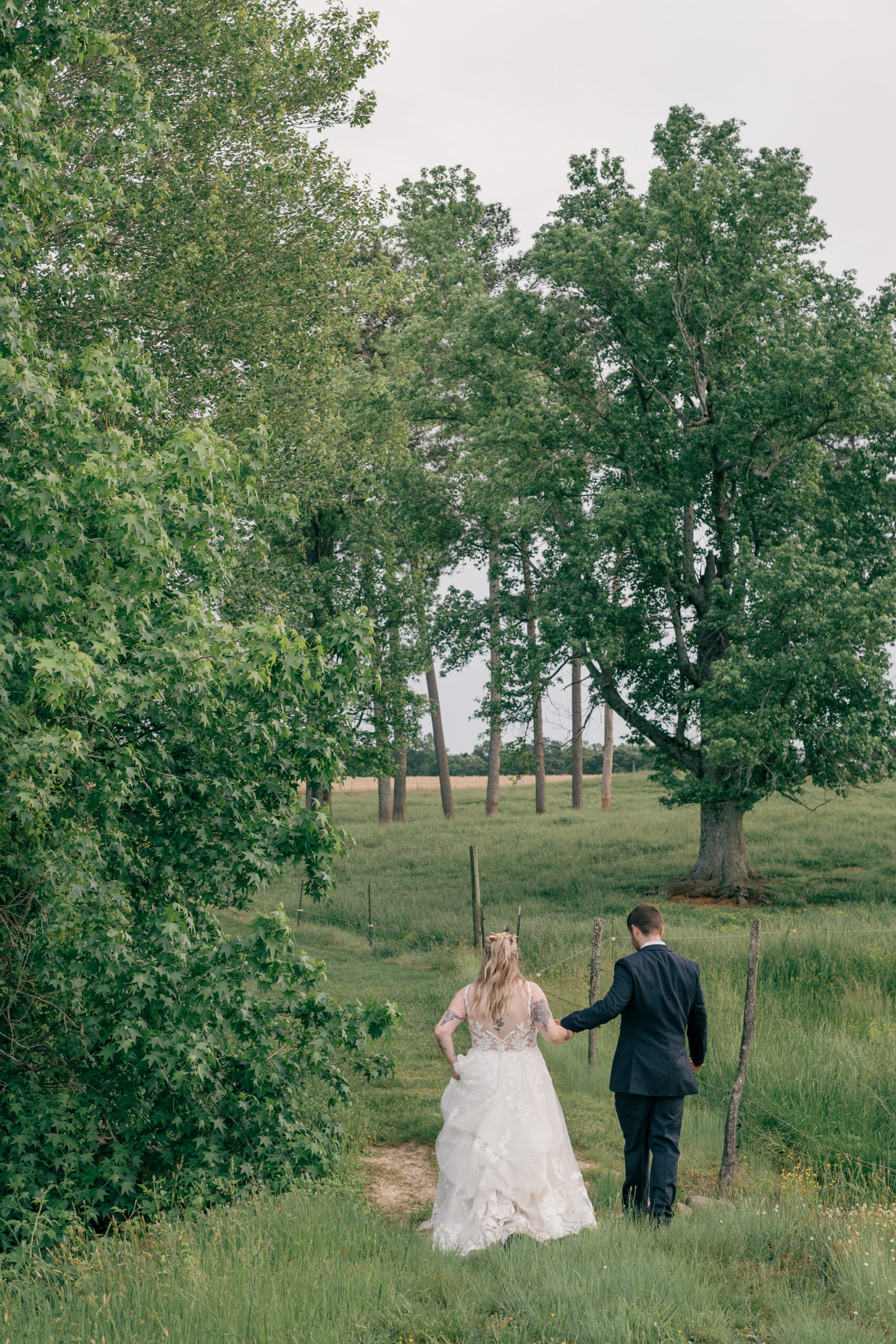 Southern MD Wedding Photographer | Bride and groom walking hand in hand down a grassy path surrounded by tall trees and open fields.