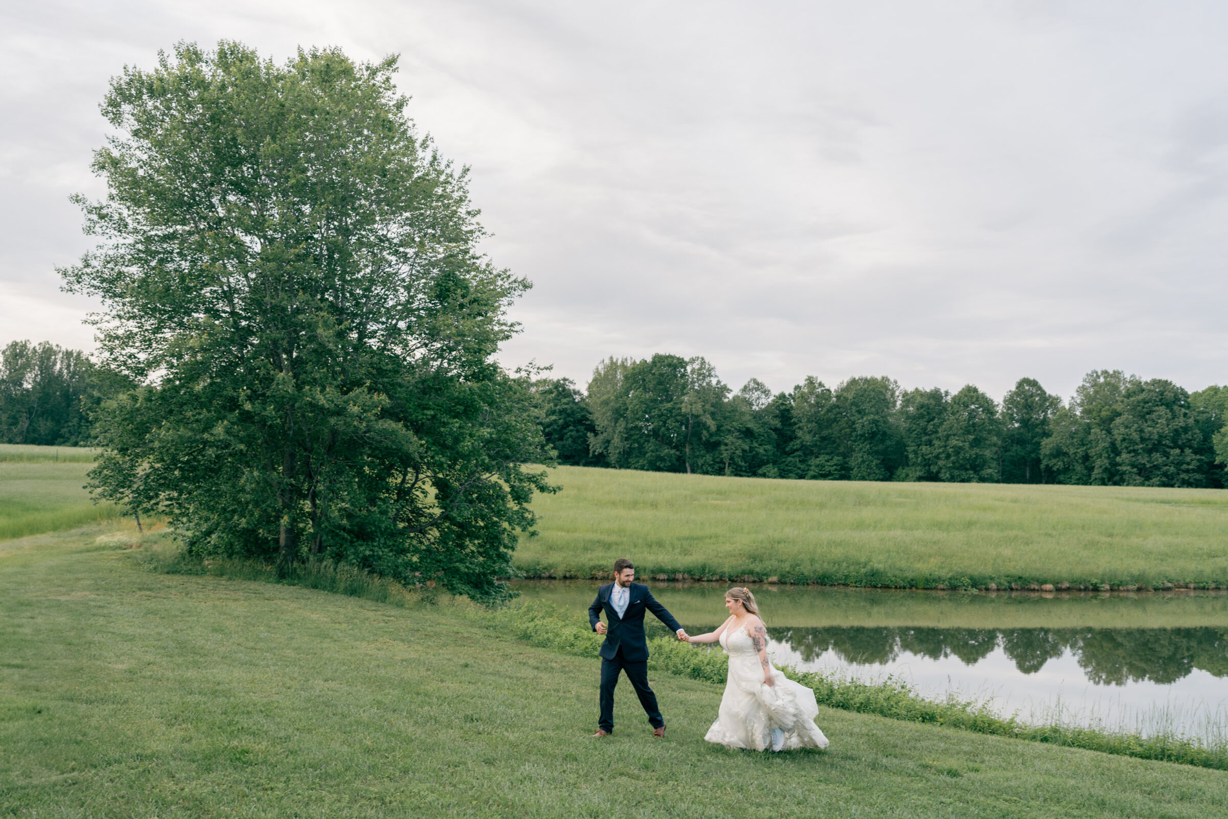 Southern Maryland Wedding Photographer | Bride and groom holding hands and running along a quiet pond, surrounded by open fields and tall trees at their countryside wedding.