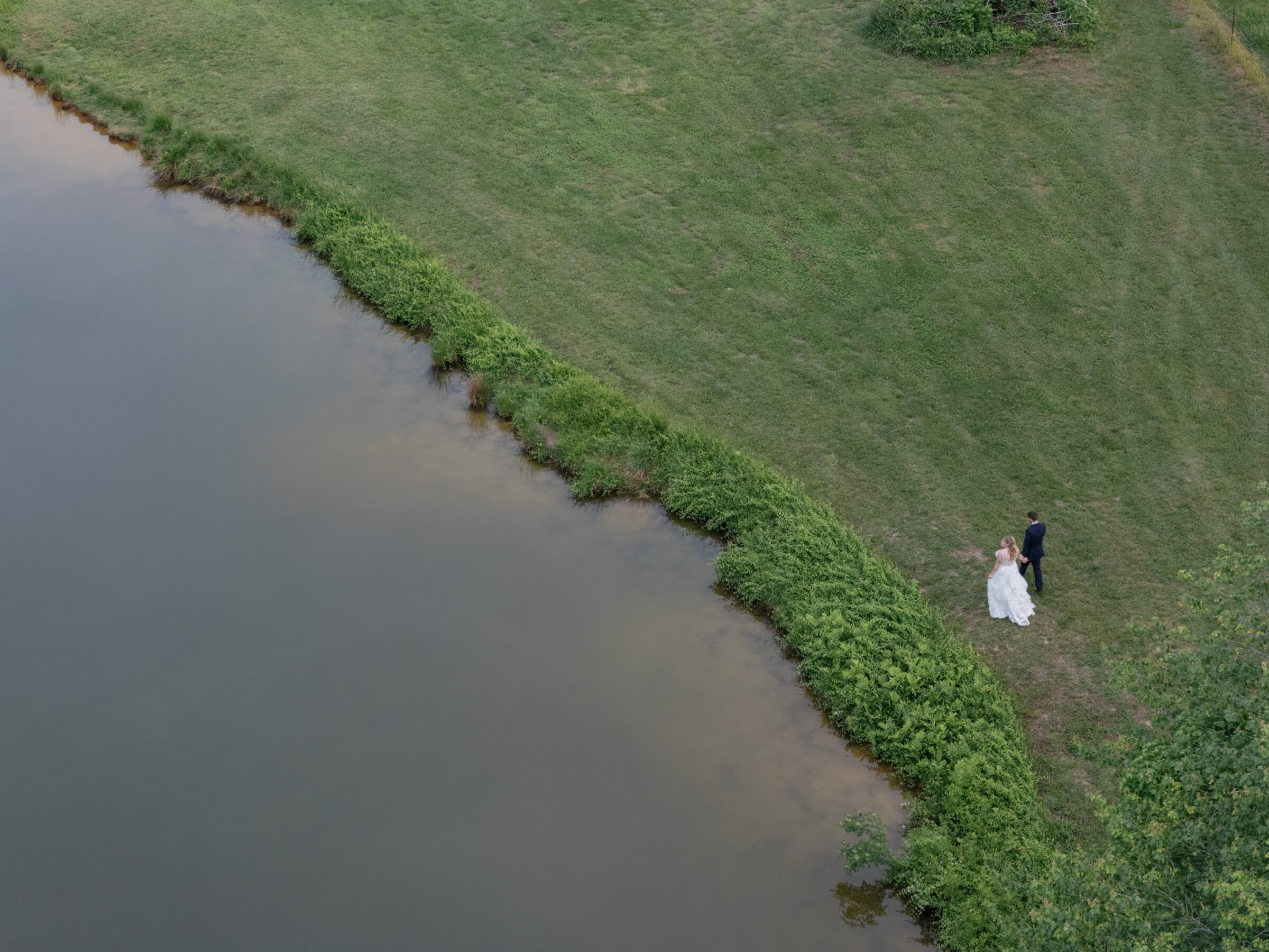 Southern Maryland Wedding Photographer | Drone view of a bride and groom walking together along the edge of a quiet pond, surrounded by open green fields.