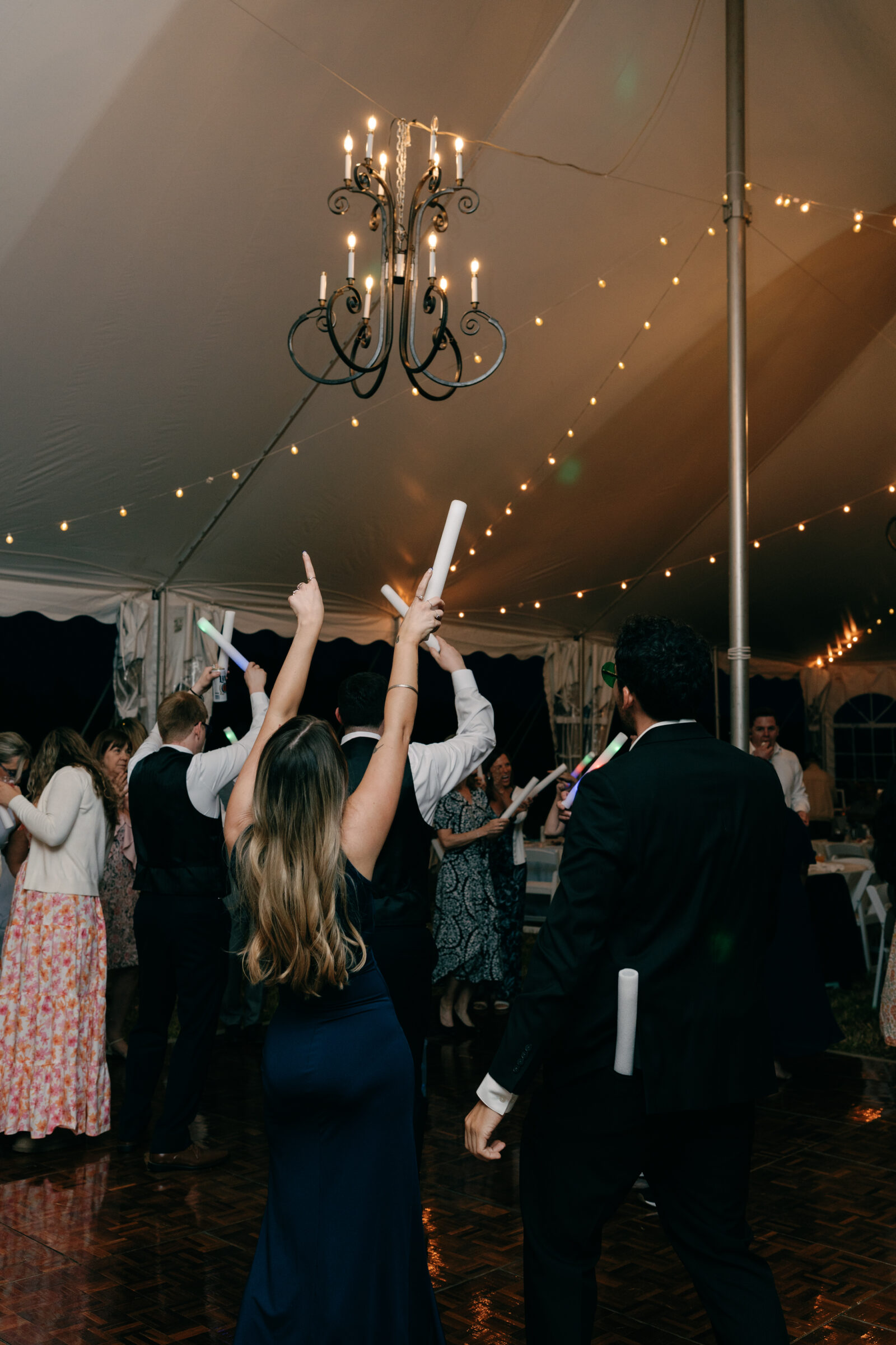 Guests dance under a tented reception with string lights and chandeliers glowing overhead, holding light-up foam sticks.