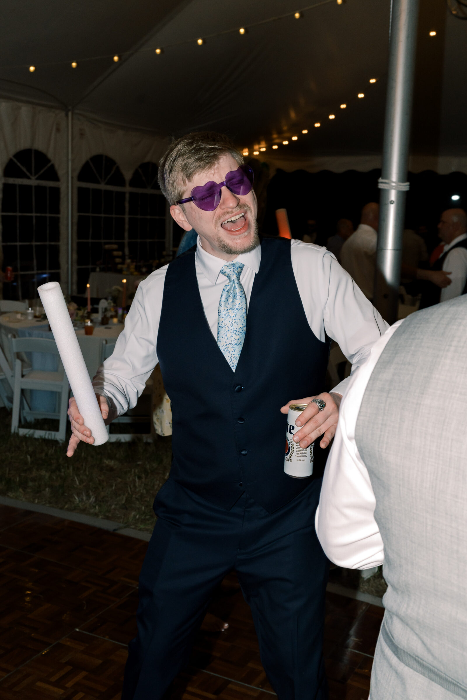 A wedding guest wearing purple heart-shaped sunglasses dances under the tented reception lights, holding a drink and a foam light stick.