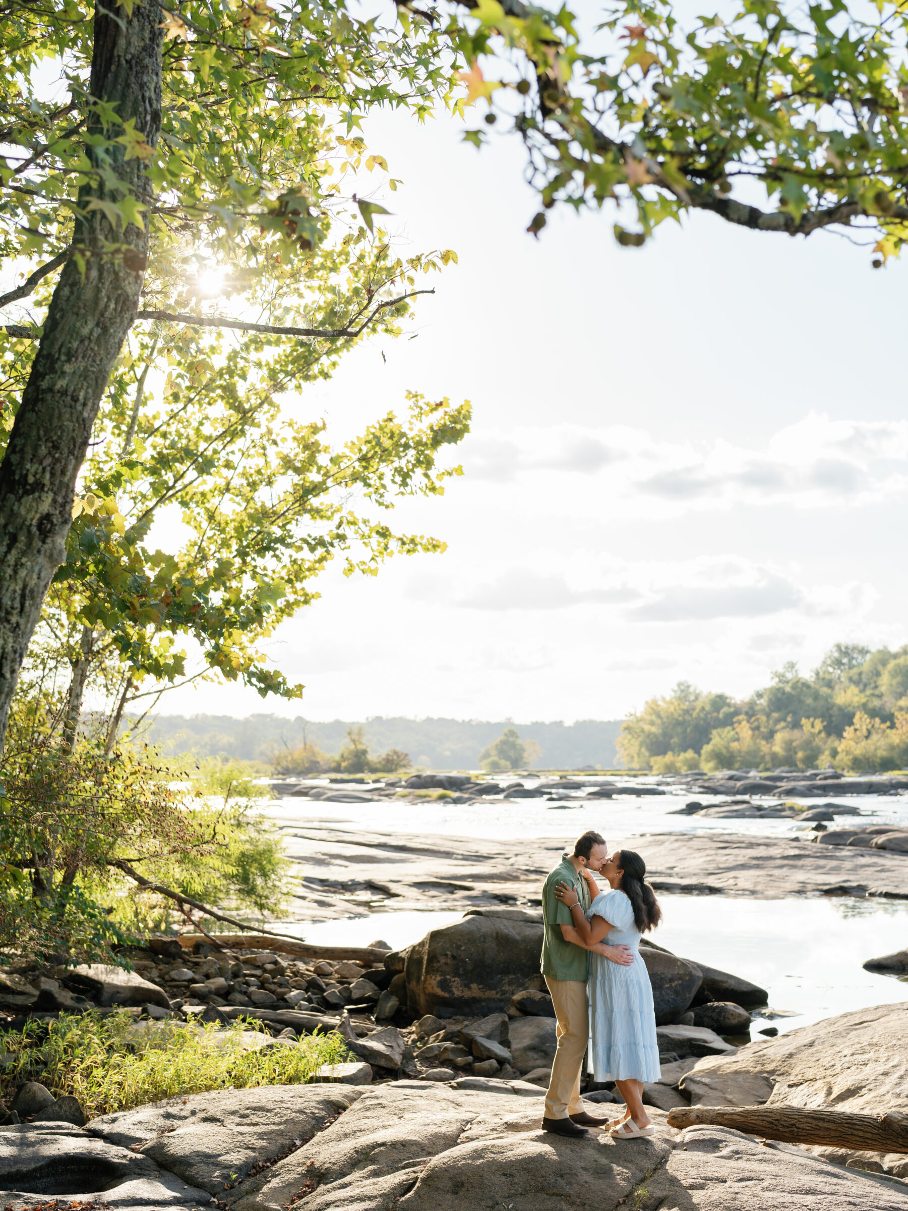 A couple shares a kiss on the rocks at Belle Isle during their Richmond engagement session, surrounded by sunlit trees and the James River’s glowing, early autumn light.