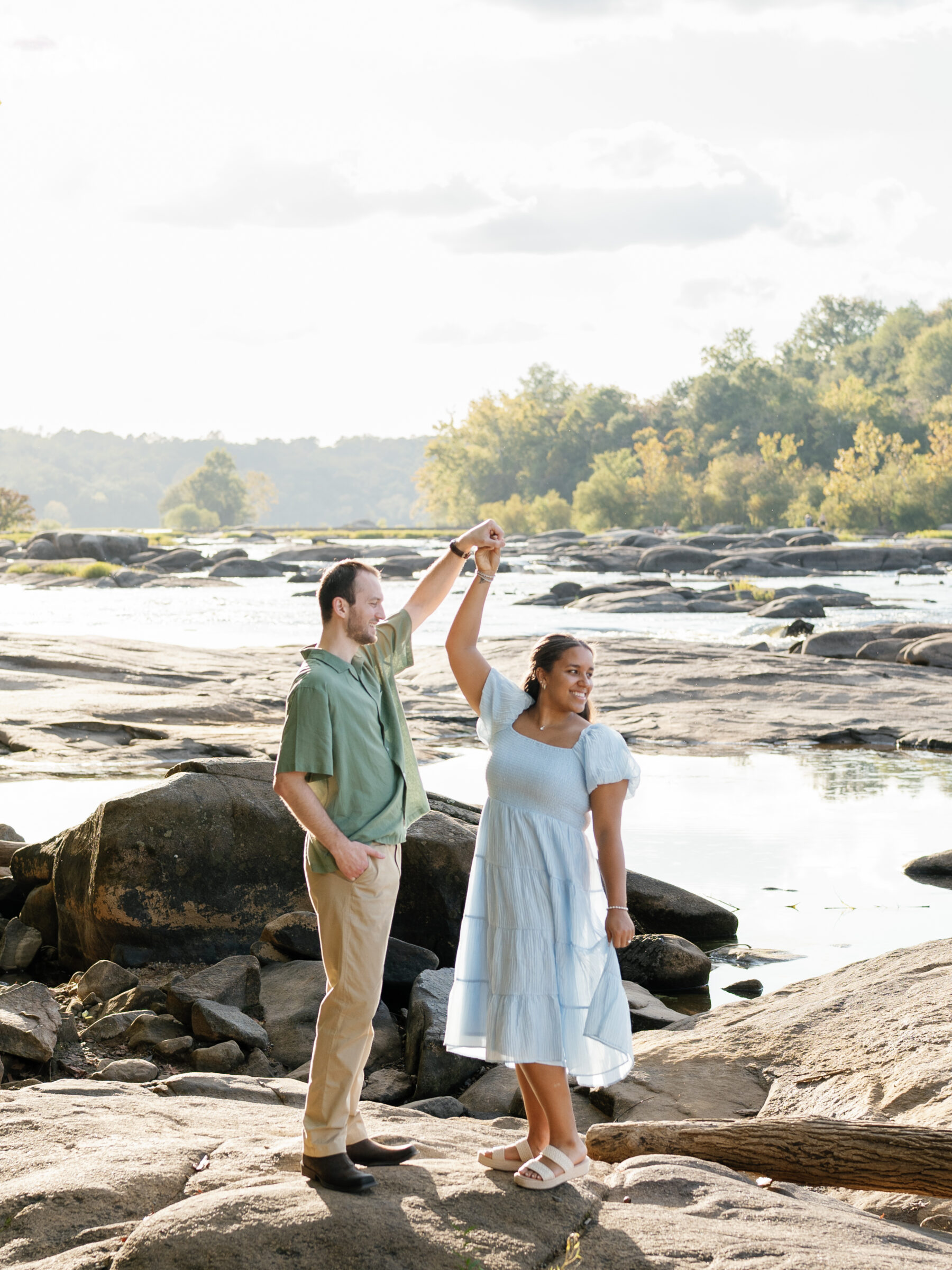 A couple dances on the sunlit rocks at Belle Isle during their Richmond engagement session, smiling as they twirl beside the James River.