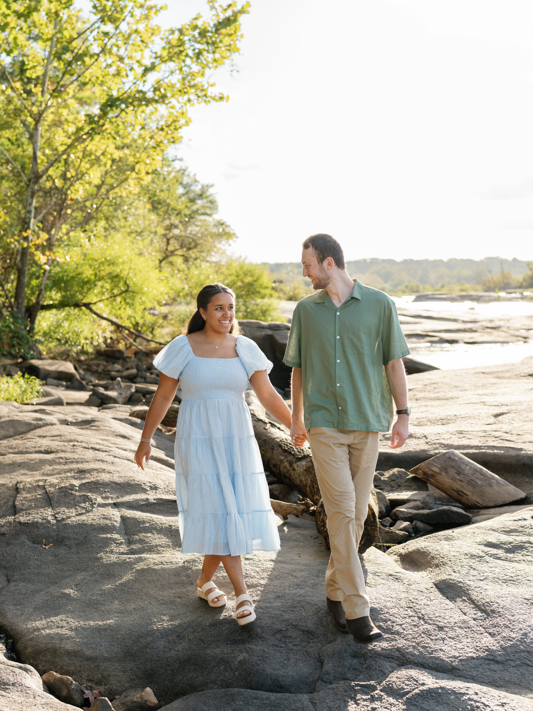 A couple walks hand in hand across the sunlit rocks at Belle Isle during their Richmond engagement session, smiling at each other in the warm early-autumn light.