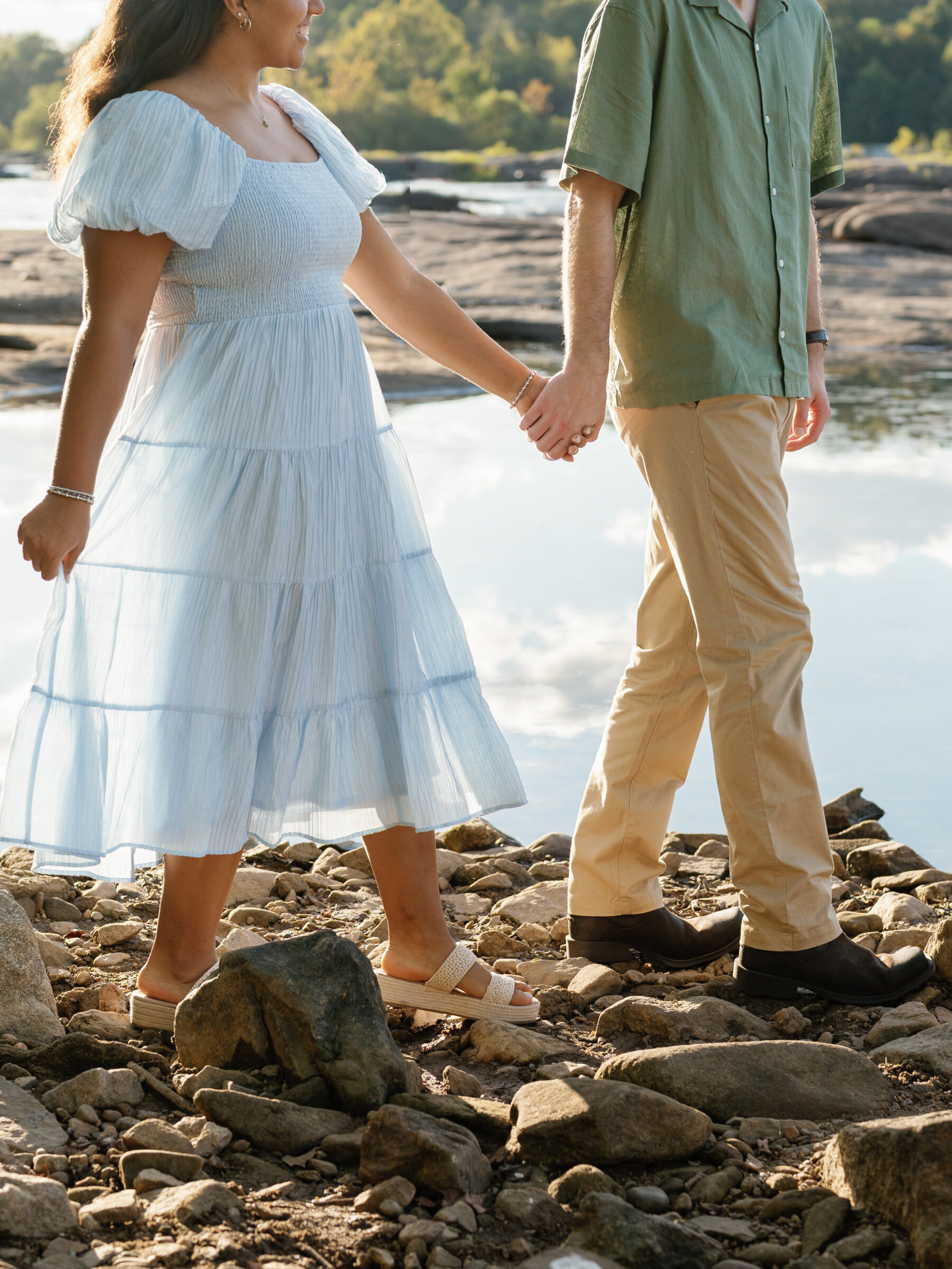 A close-up of a couple holding hands as they walk along the rocky shoreline at Belle Isle during their Richmond engagement session, sunlight catching the soft blue of her dress.