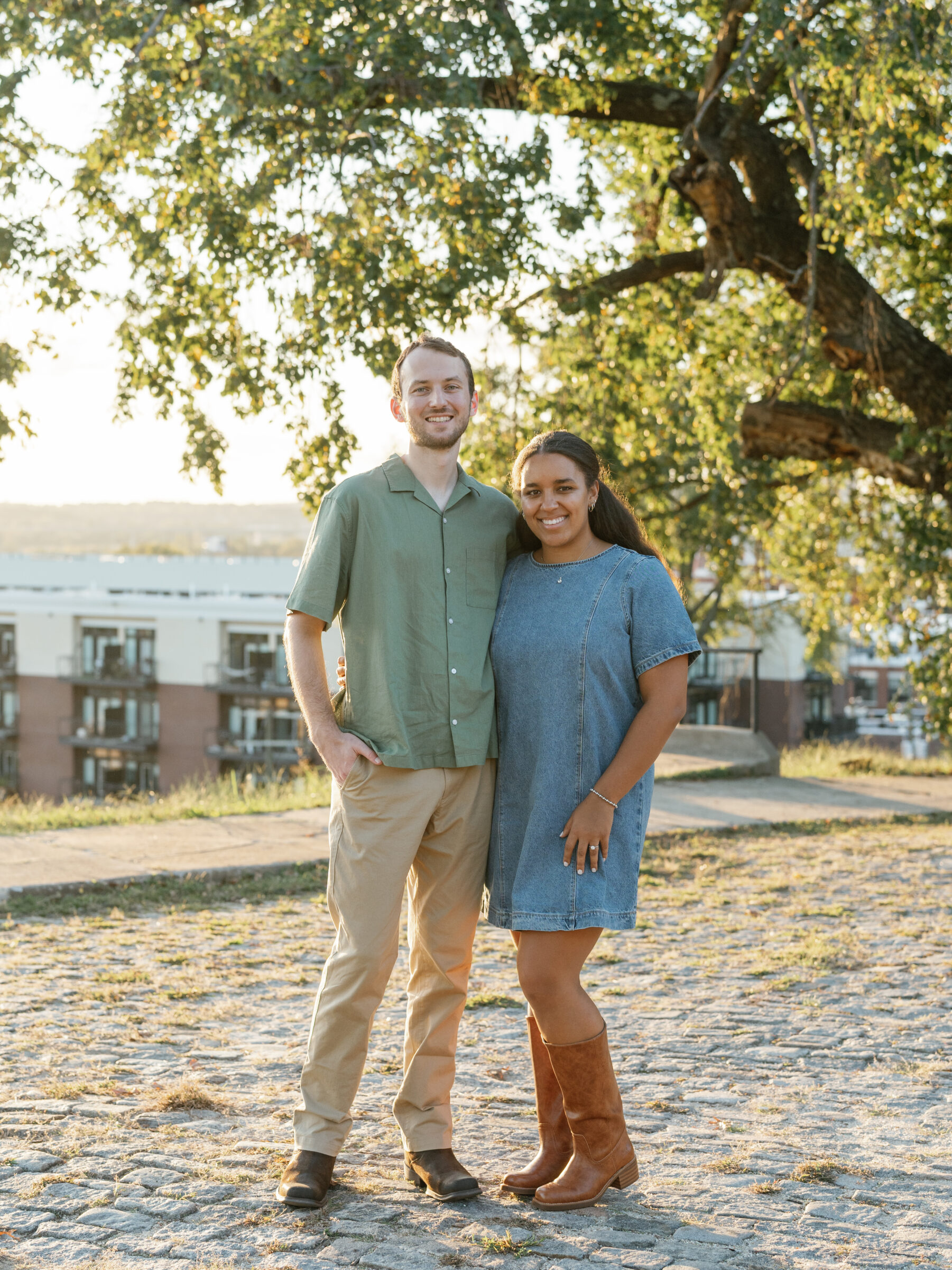 A couple stands together at Libby Hill Park during their Richmond engagement session, smiling in the warm sunset light beneath a large, leafy tree.
