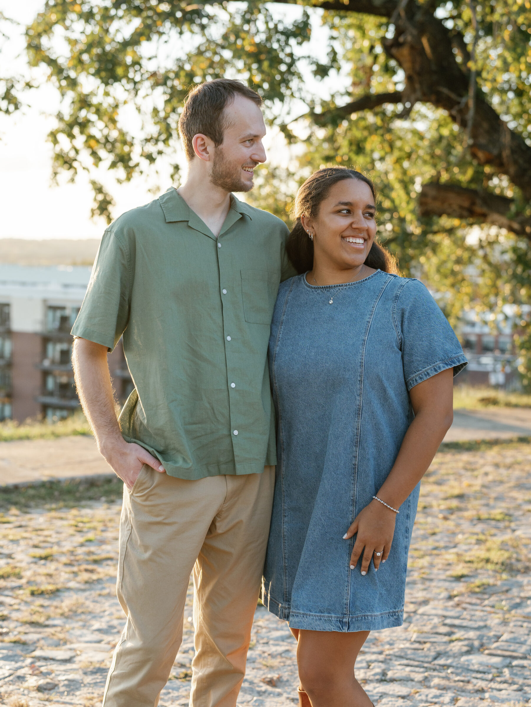 A couple smiles together at Libby Hill Park during their Richmond engagement session, standing close in the warm golden-hour light beneath a leafy tree.