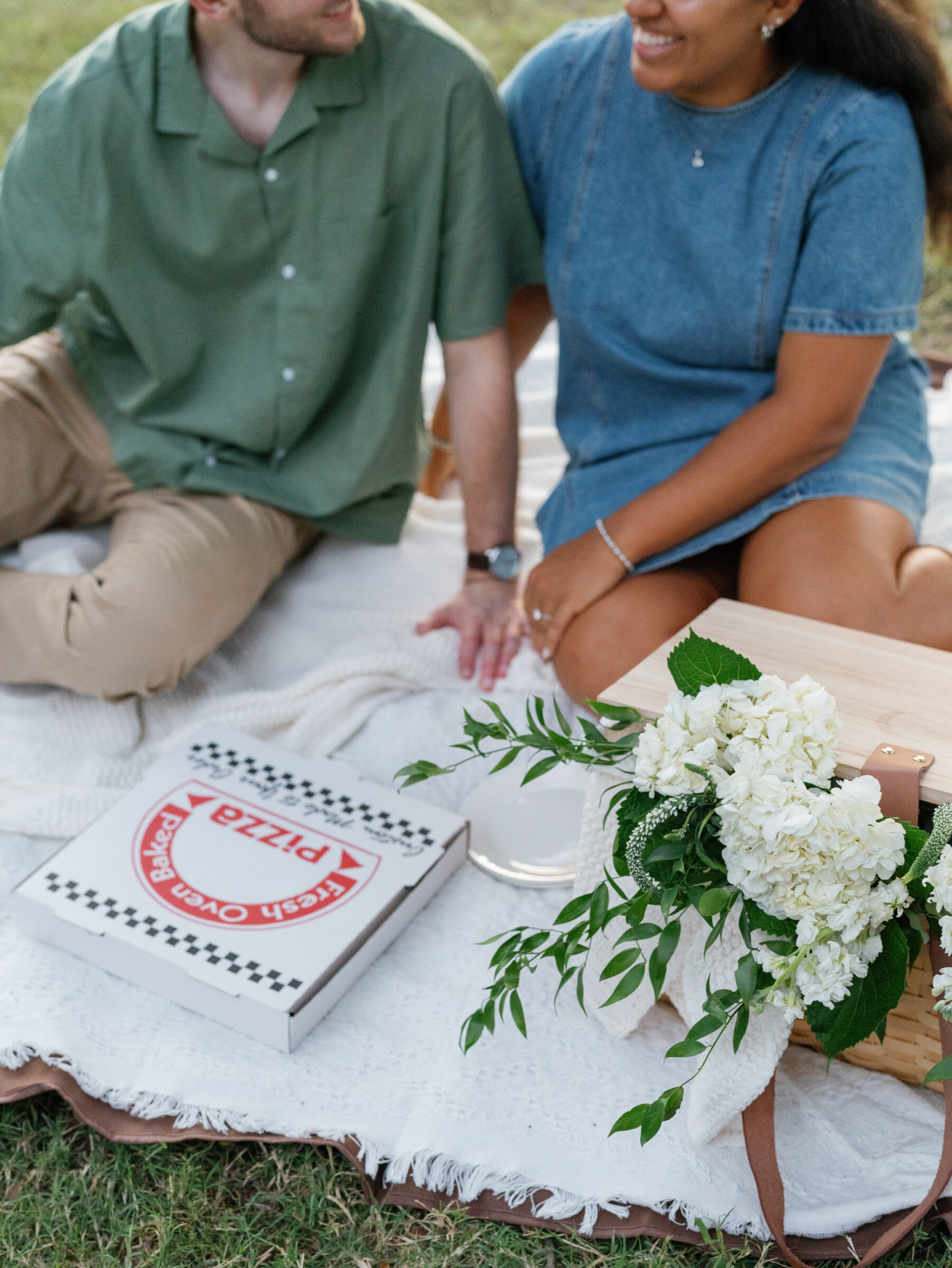 A couple sits together during their pizza picnic at Libby Hill Park, with a fresh pizza box, a woven basket, and white flowers arranged on a blanket.