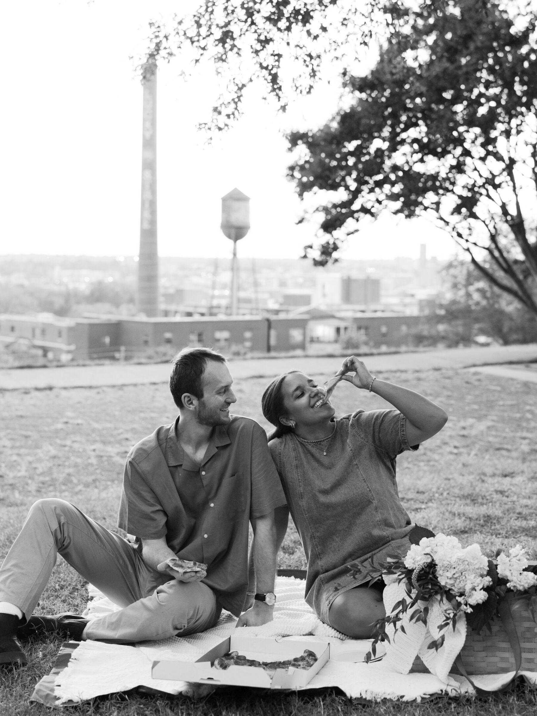 A couple enjoys a playful pizza picnic at Libby Hill Park during their Richmond engagement session, laughing together as she takes a bite of pizza.