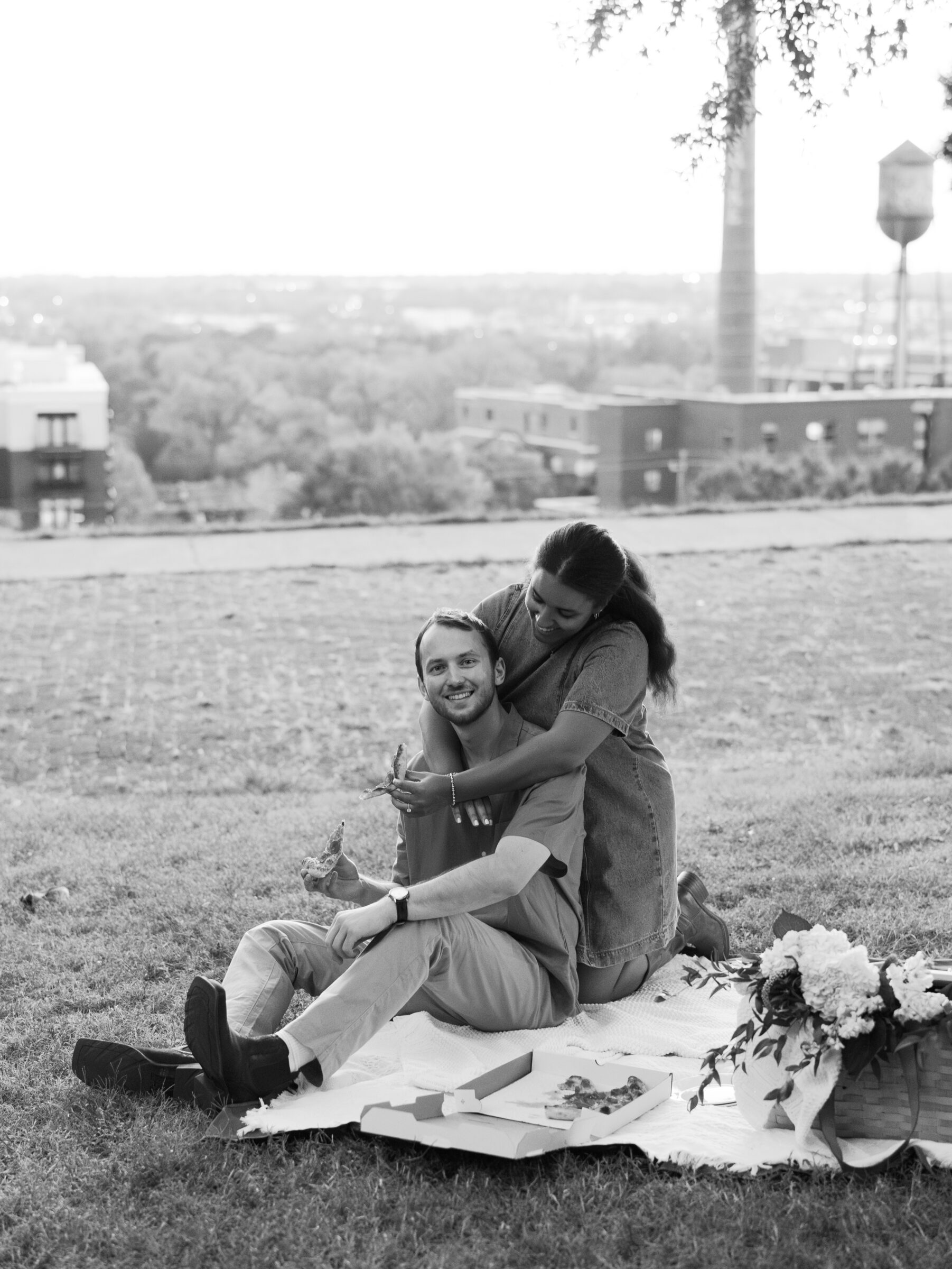 A couple shares a playful moment during their pizza picnic at Libby Hill Park, with she wrapping her arms around him from behind as he smiles at the camera.