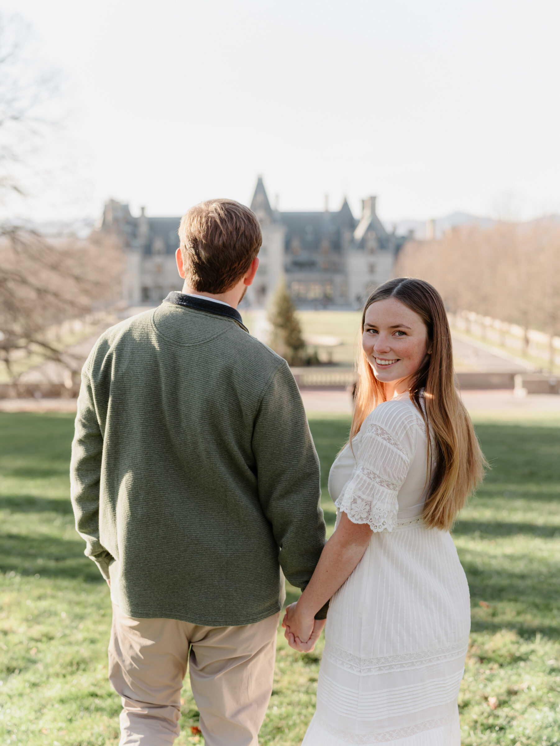 Bride looking back toward the camera while holding hands with her fiancé at the Biltmore Estate during a fall engagement session in Asheville NC