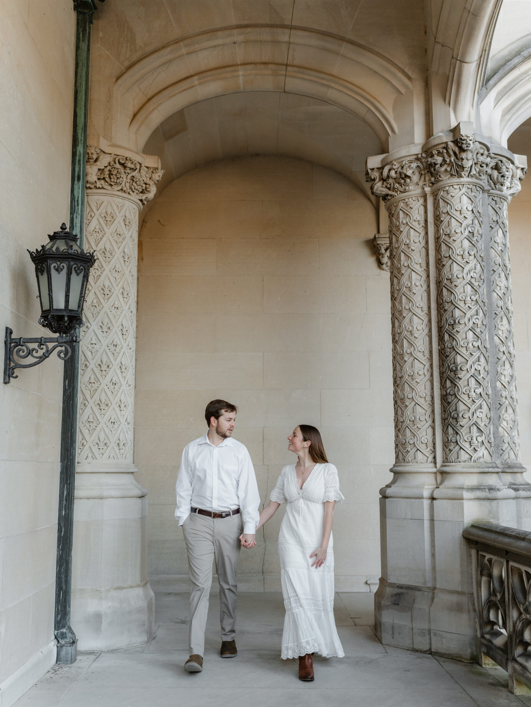 Couple walking hand in hand beneath the stone arches at the Biltmore Estate during an editorial engagement session in Asheville NC