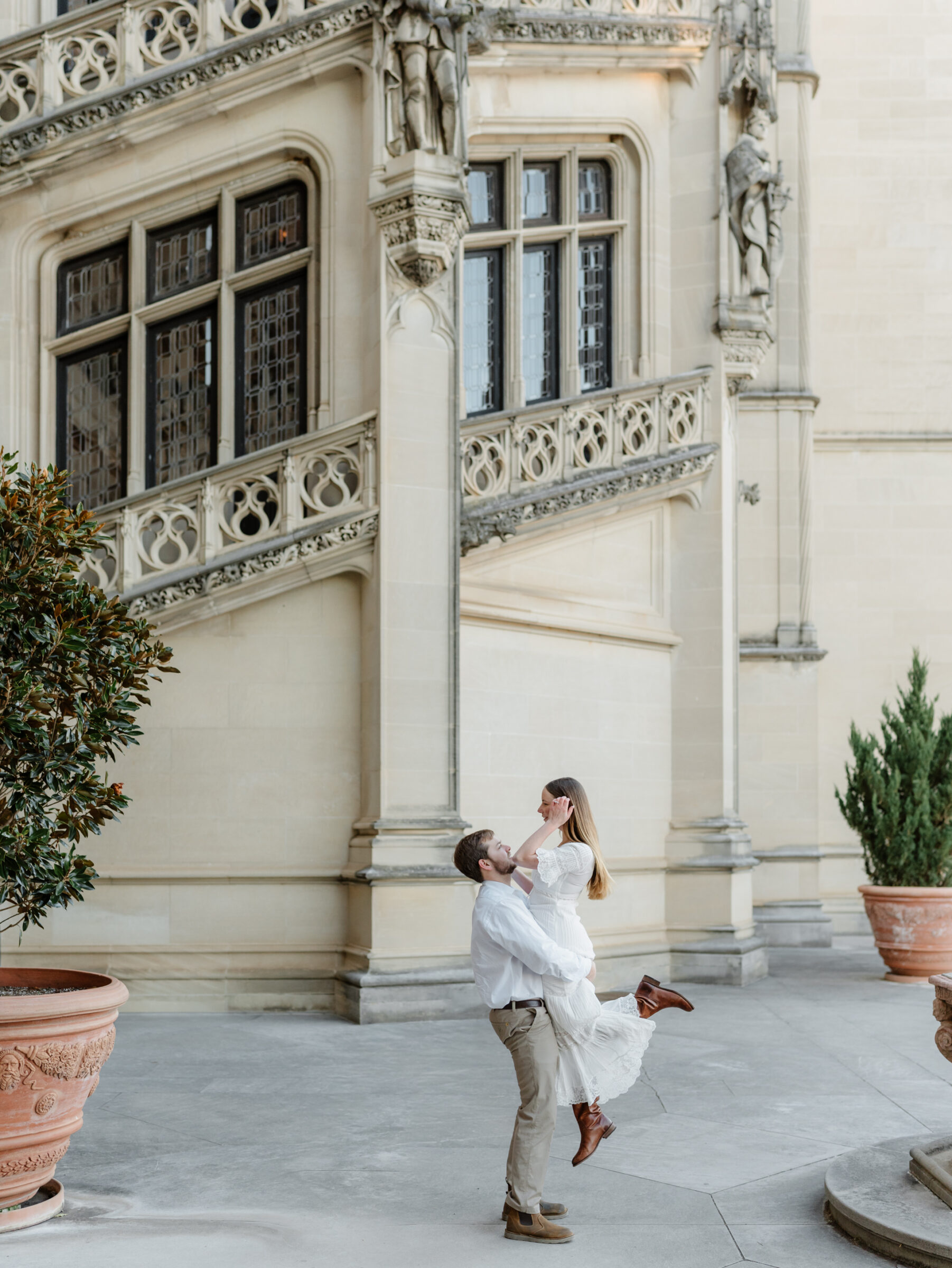 Candid engagement moment of a groom lifting his partner in front of historic stone architecture