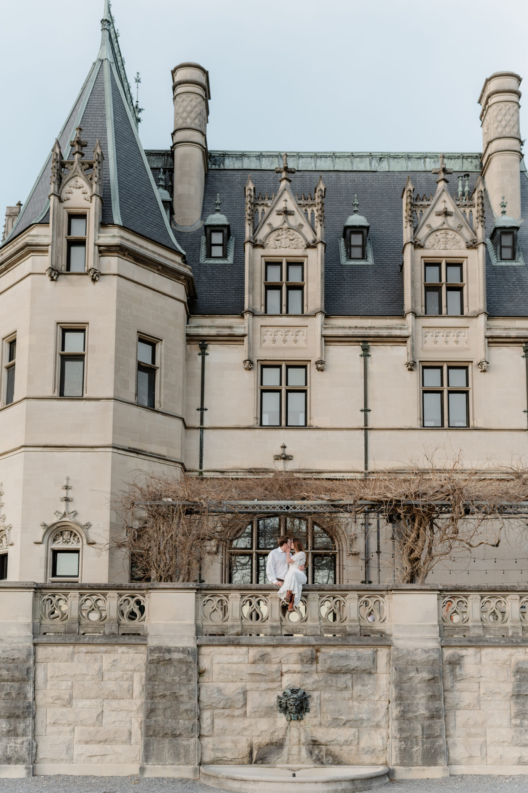Wide architectural portrait of a couple sharing an intimate moment on a stone terrace beneath a grand historic estate