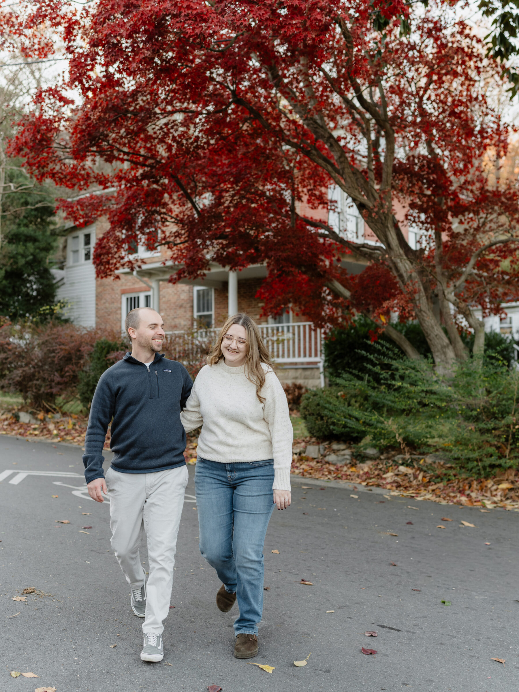 Couple walking arm in arm under a bright red autumn tree in Historic Occoquan during their fall engagement session.