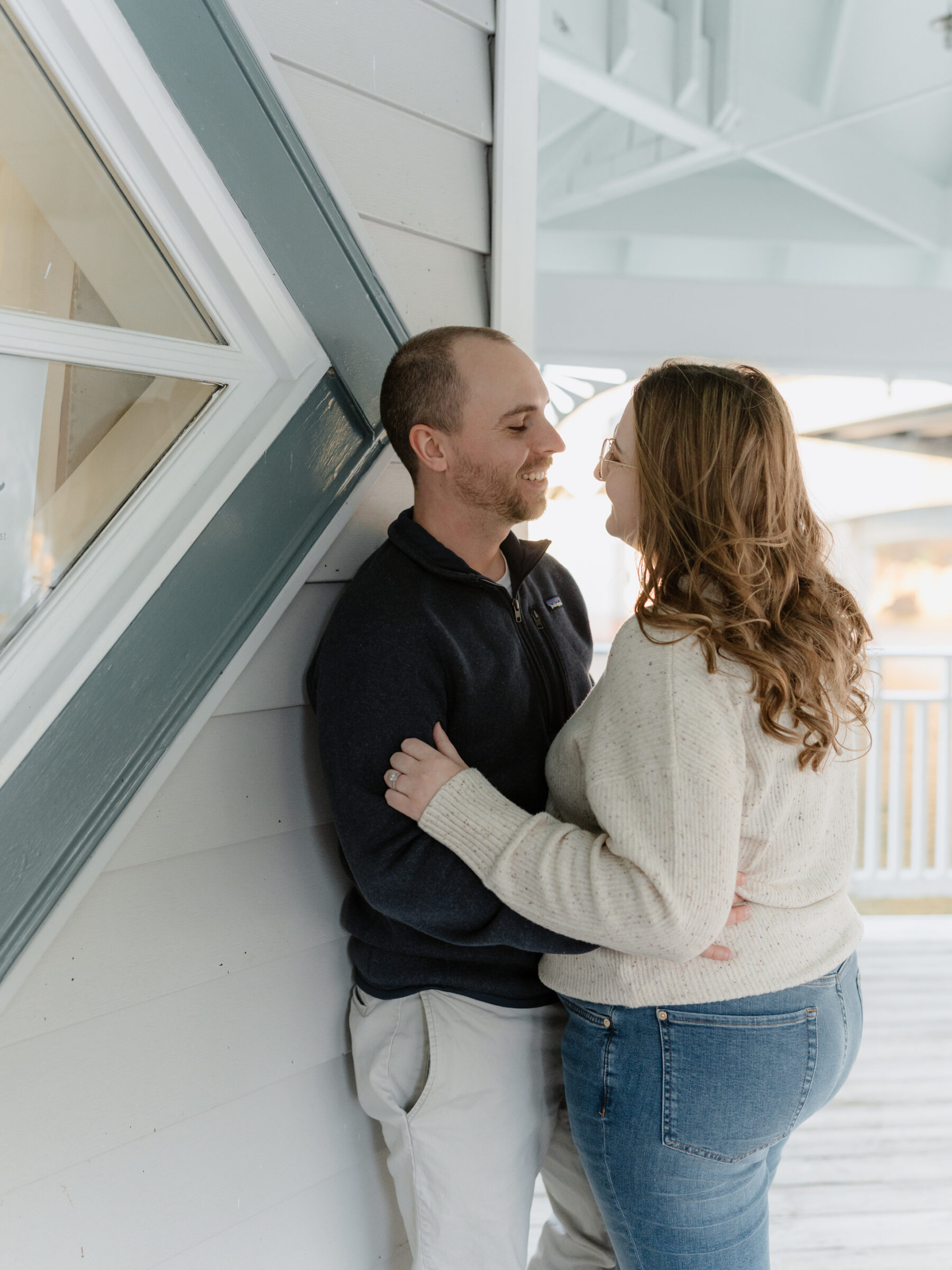 Couple standing close together on a waterfront porch, smiling at each other during their fall engagement session.
