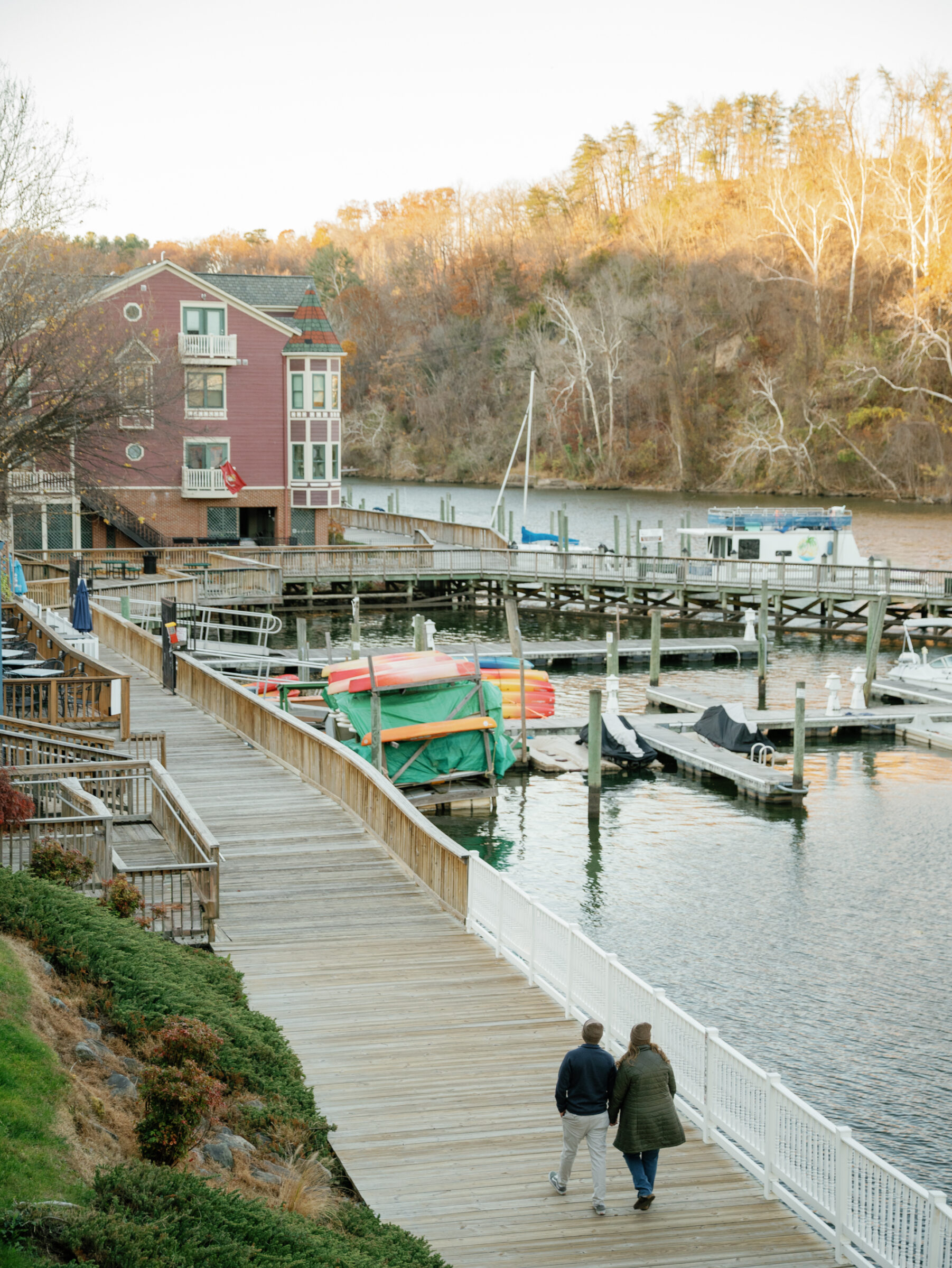 Couple walking along the wooden boardwalk in Historic Occoquan with views of the waterfront, boats, and autumn trees.