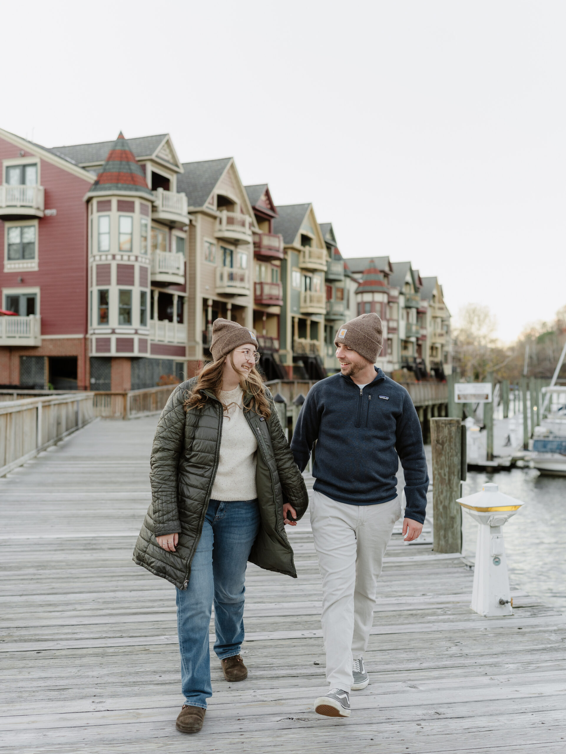 Couple walking hand in hand along the wooden dock in Historic Occoquan with colorful waterfront buildings in the background.