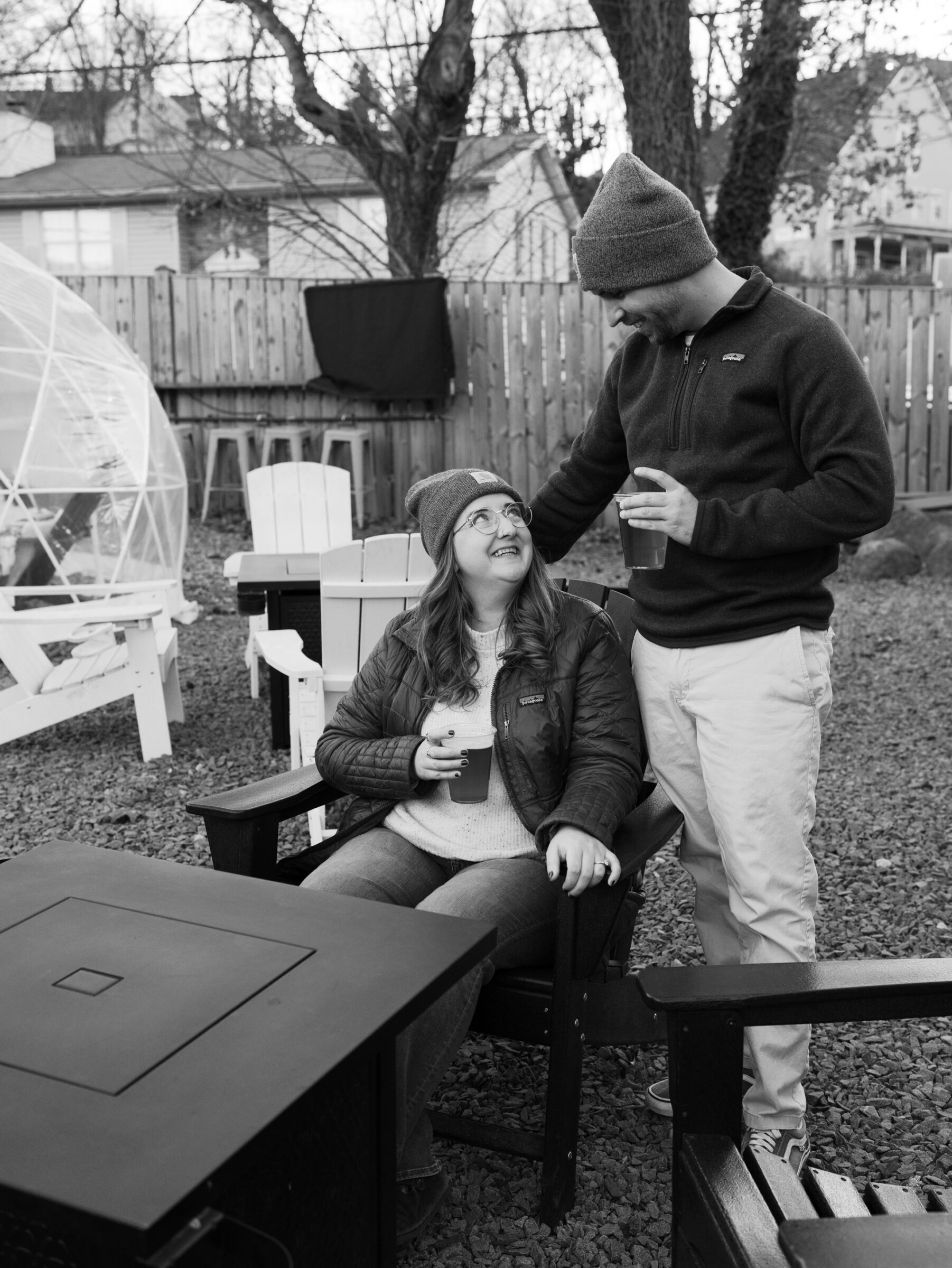 Couple enjoying drinks together around a fire pit, with the woman seated and looking up at her partner.