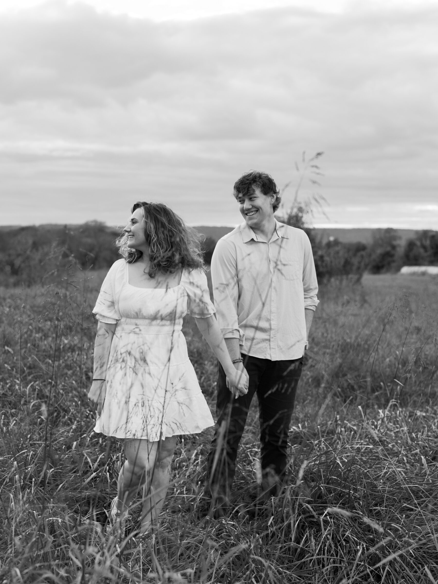 Lovettsville VA Wedding Photographer | Black and white engagement photo of a couple walking hand in hand through tall grass during an at-home session in Lovettsville, Virginia