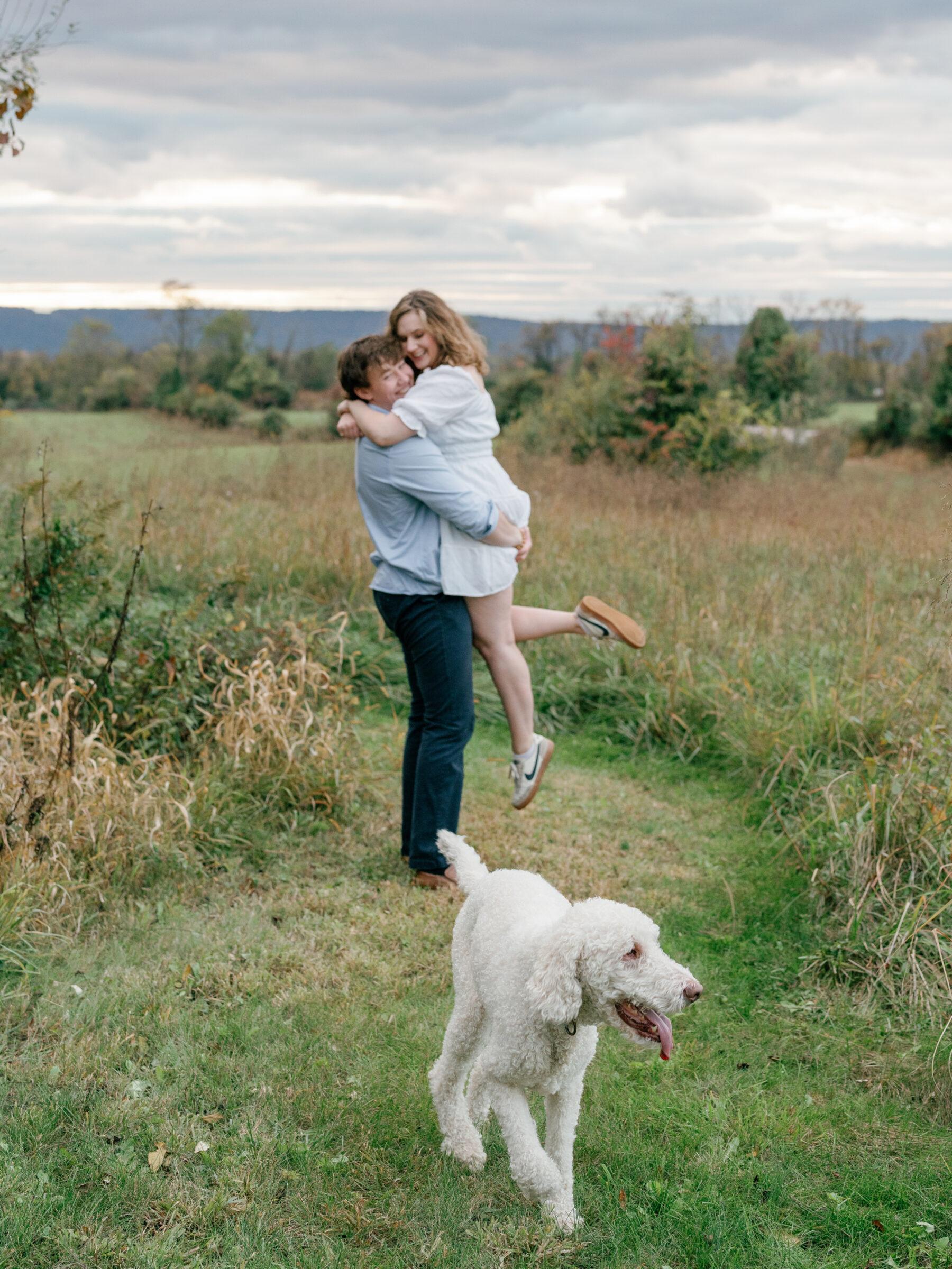 Lovettsville VA Wedding Photographer | Engaged couple laughing and embracing in a field while their dog runs ahead during an at-home engagement session in Lovettsville, Virginia