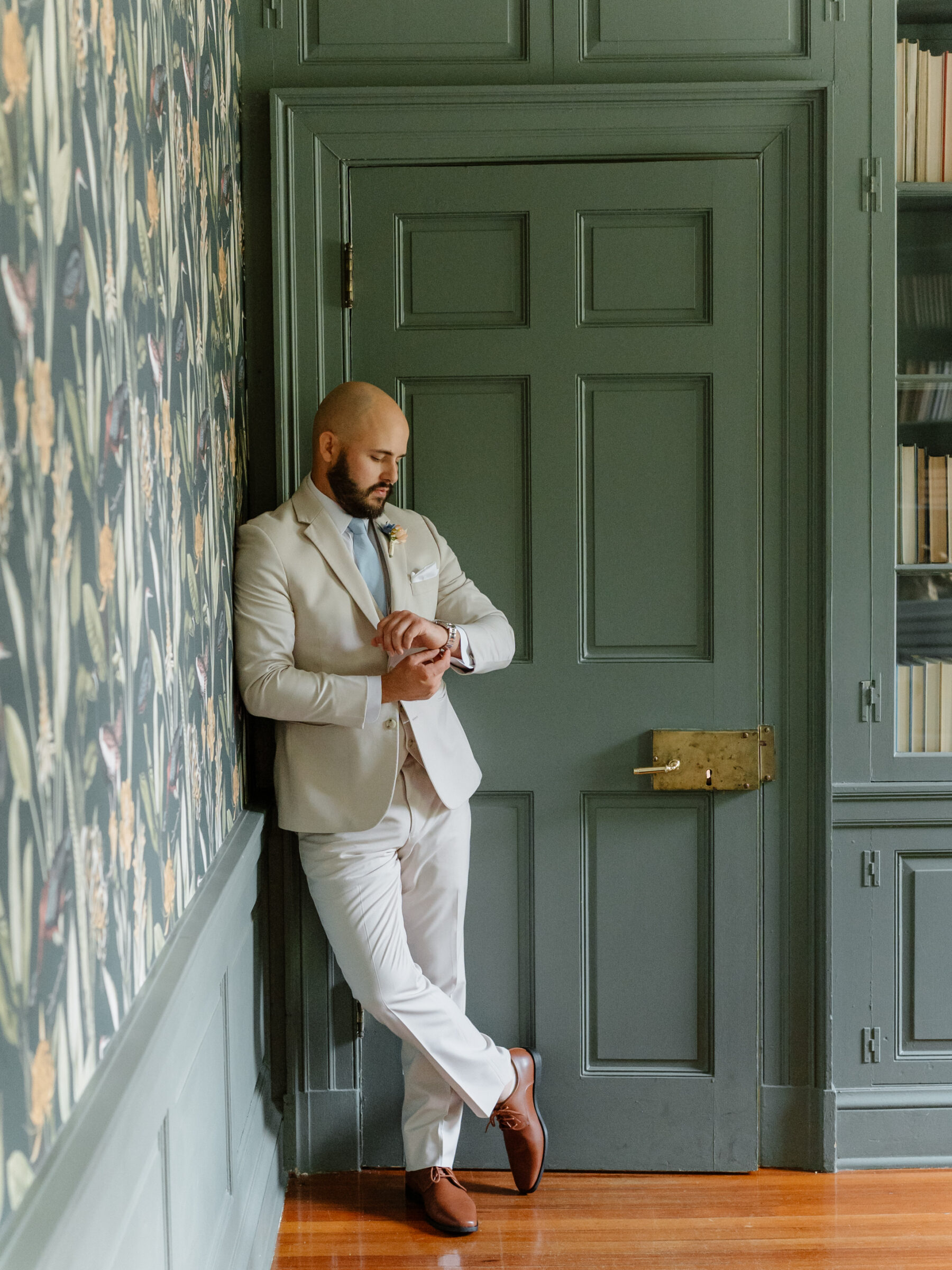 Full-length portrait of the groom standing in the manor house library, checking his watch before the wedding.