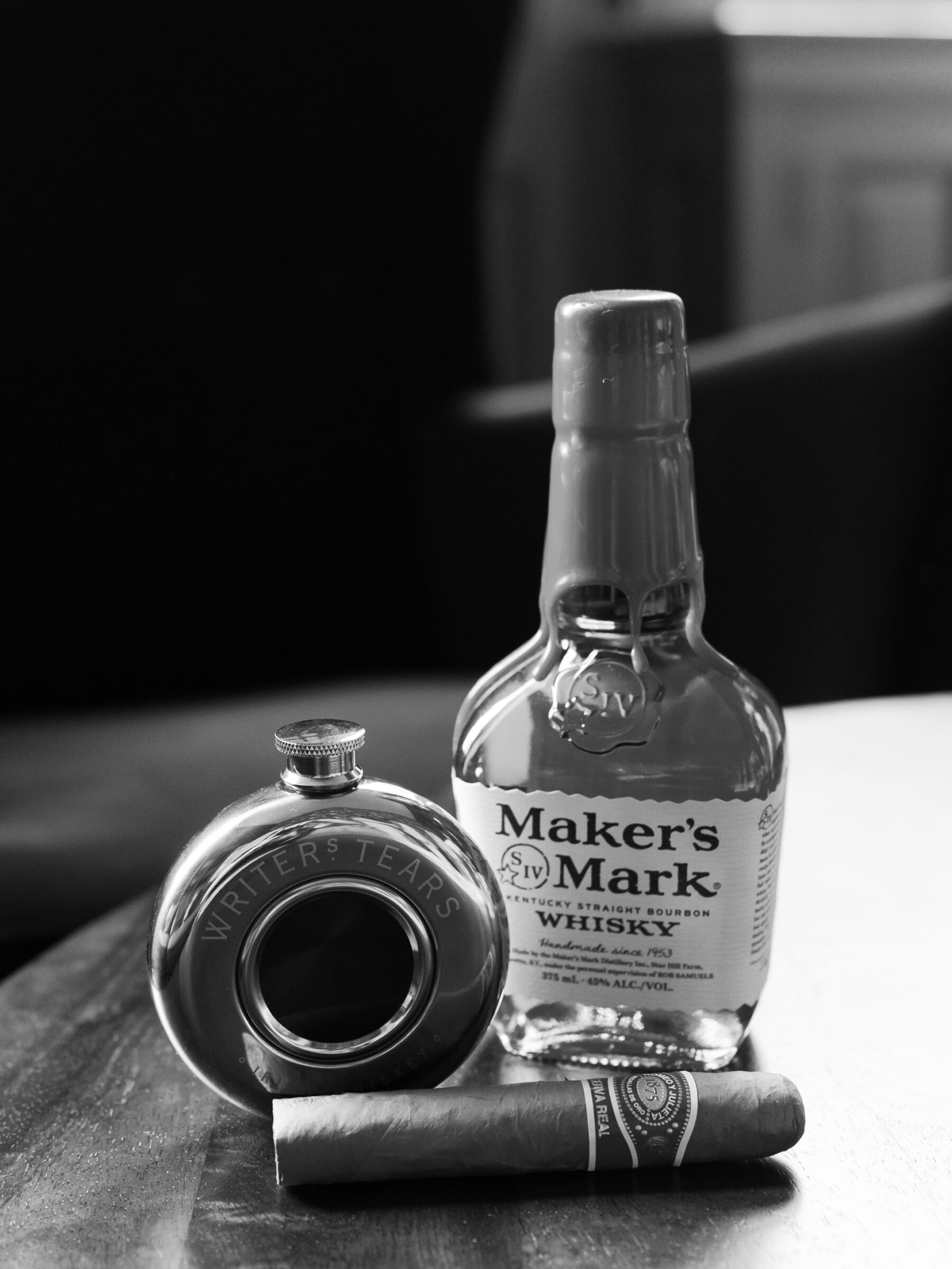 Black and white detail photo of a flask, bourbon bottle, and cigar set out before the wedding.