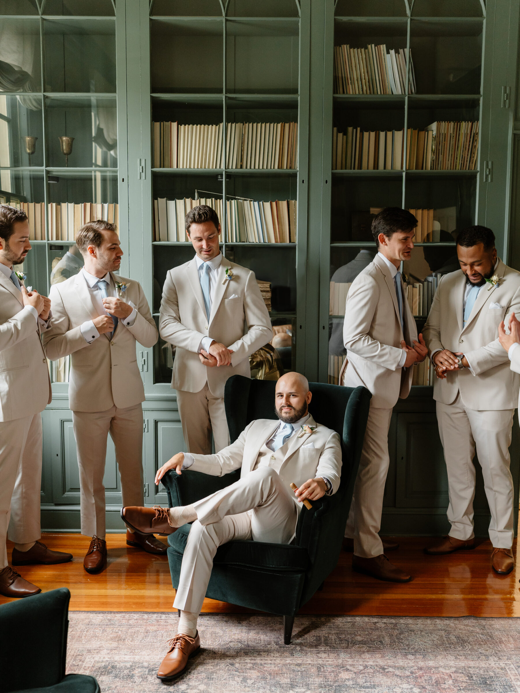 Groom seated in a green armchair with his groomsmen gathered around him inside the manor house library before the wedding.