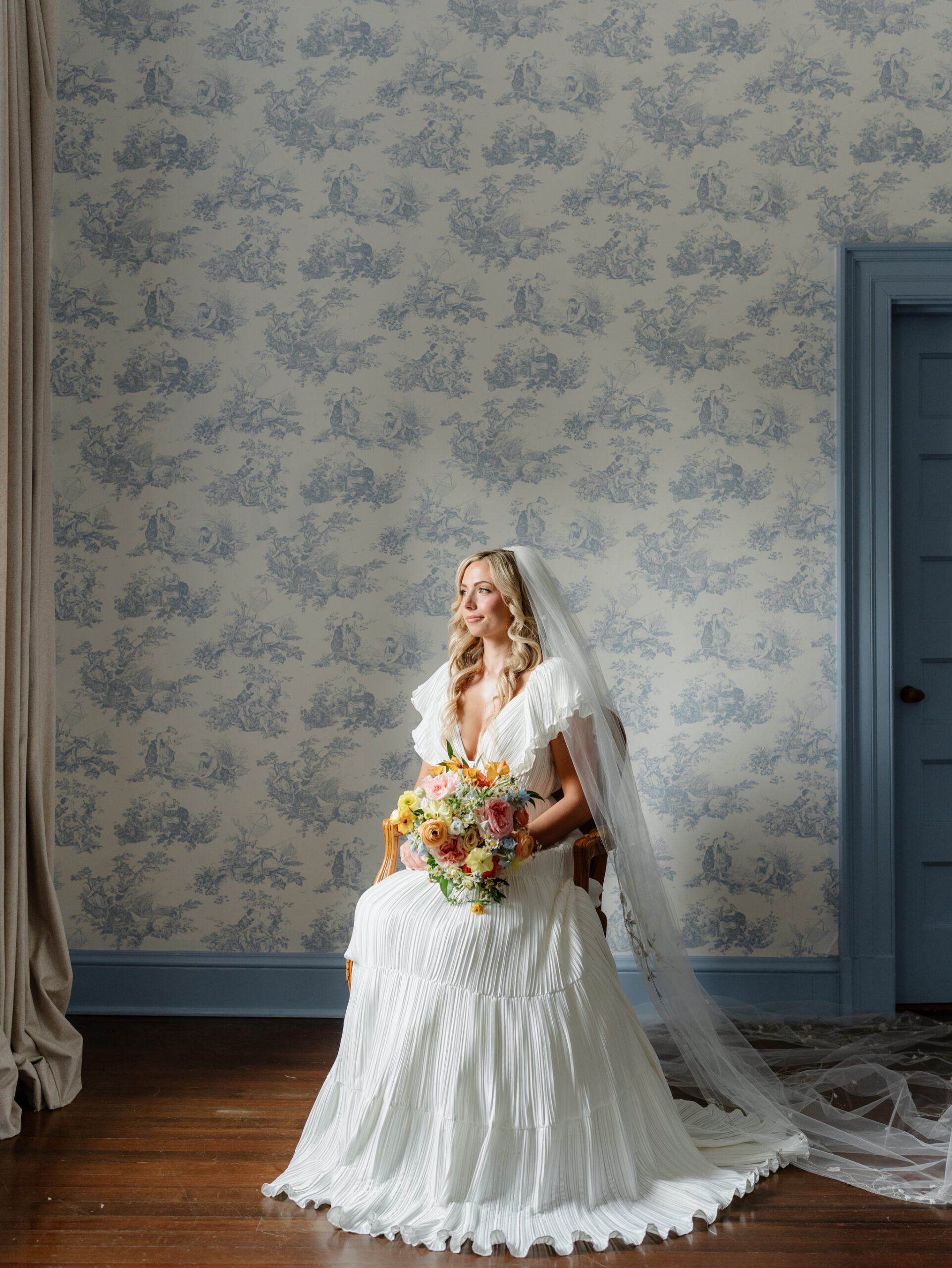 Bride seated in the Rust Manor House bridal suite holding her bouquet, framed by soft window light and blue toile wallpaper.