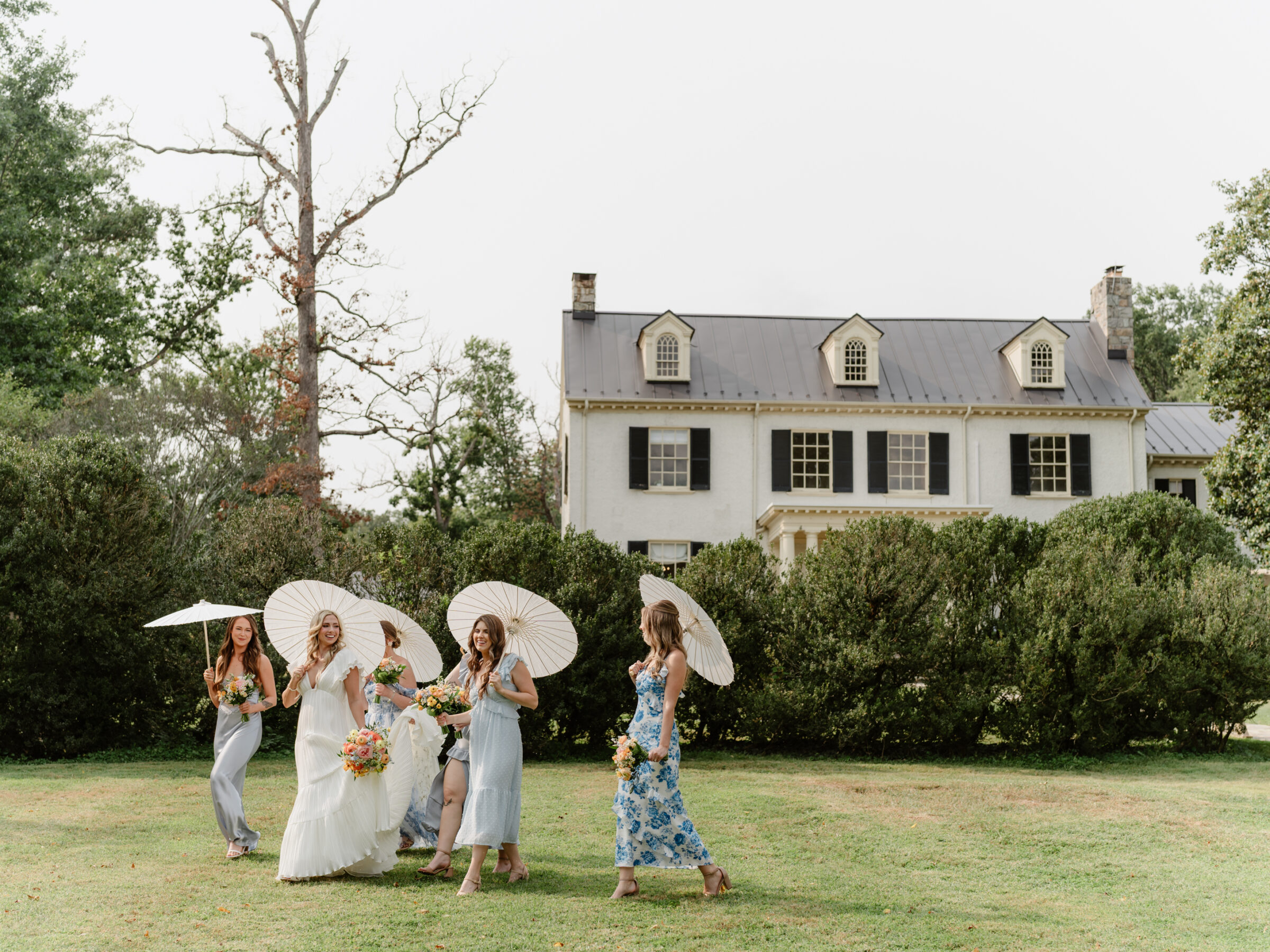 Bride walking across the lawn at Rust Manor House with her bridesmaids holding parasols before the ceremony.