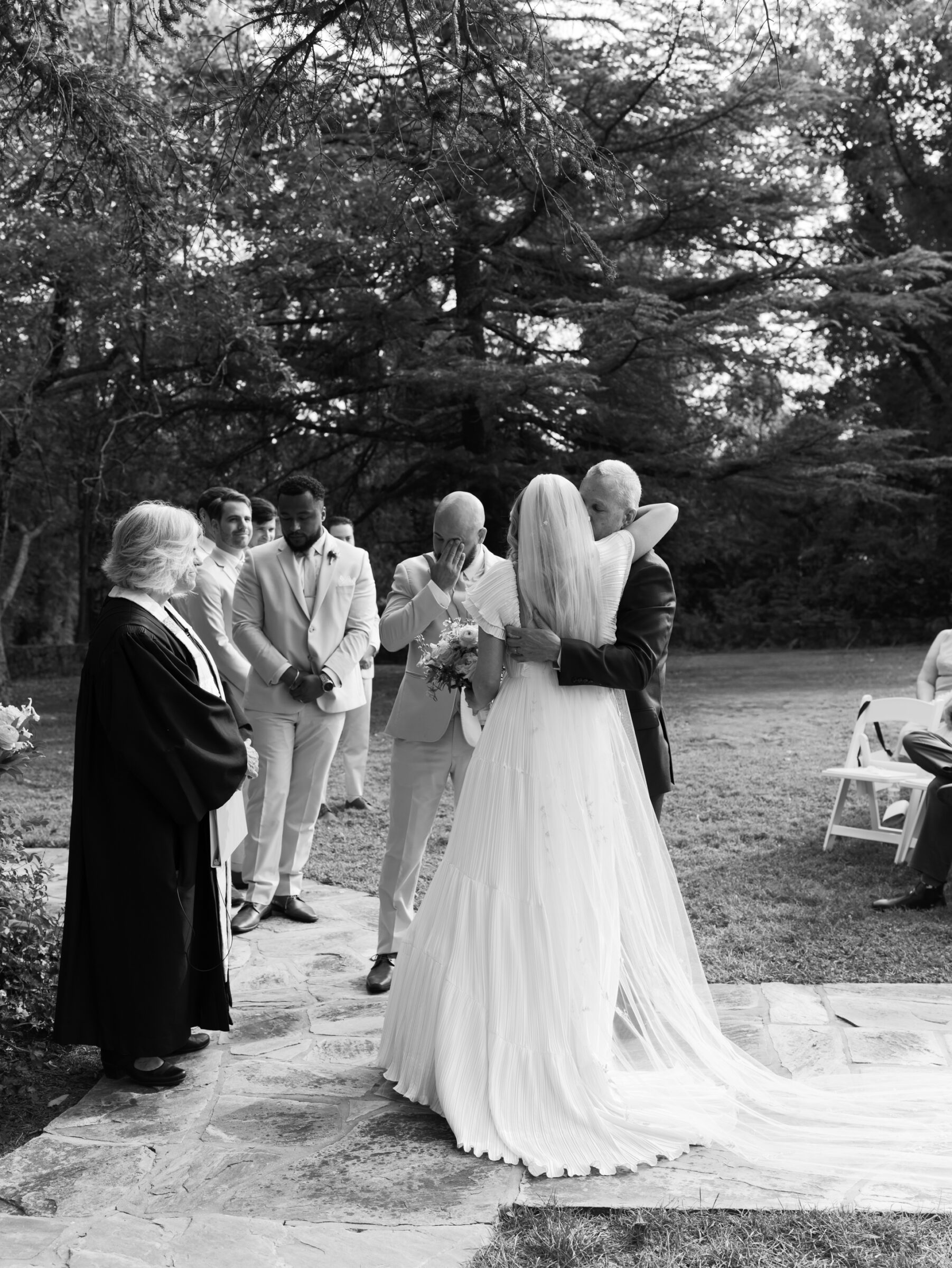 Black and white photo of the bride hugging her escort at the start of the Rust Manor House ceremony as the groom wipes away a tear.