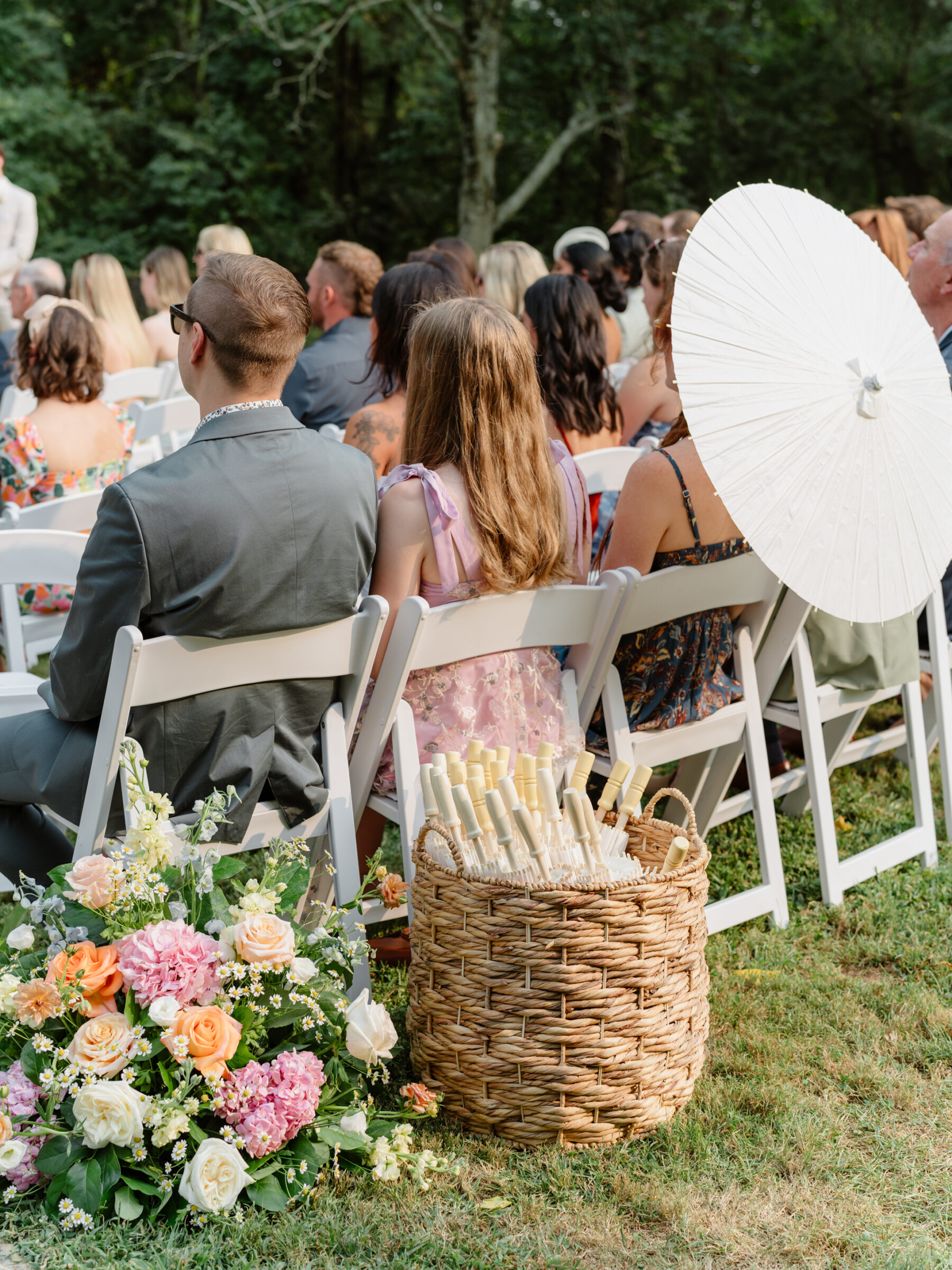 Guests seated during the garden ceremony with parasols and a basket of fans beside colorful floral arrangements.