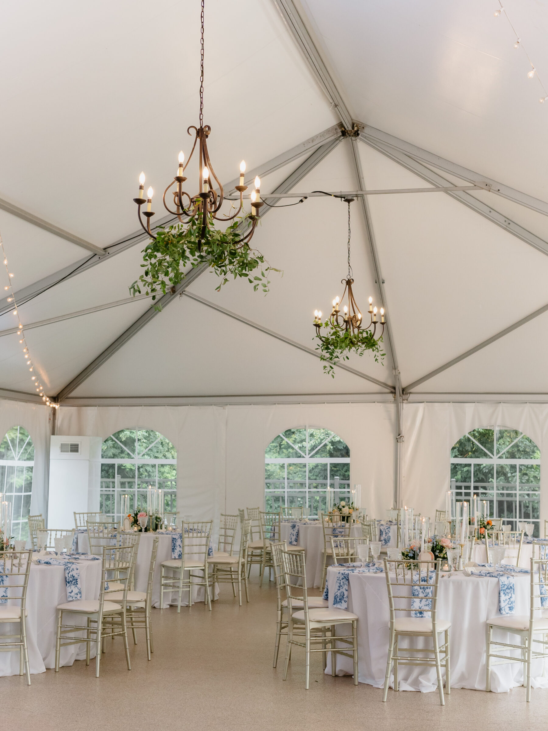 The tented reception space at Rust Manor House set with round tables, blue patterned linens, and floral centerpieces beneath hanging chandeliers.