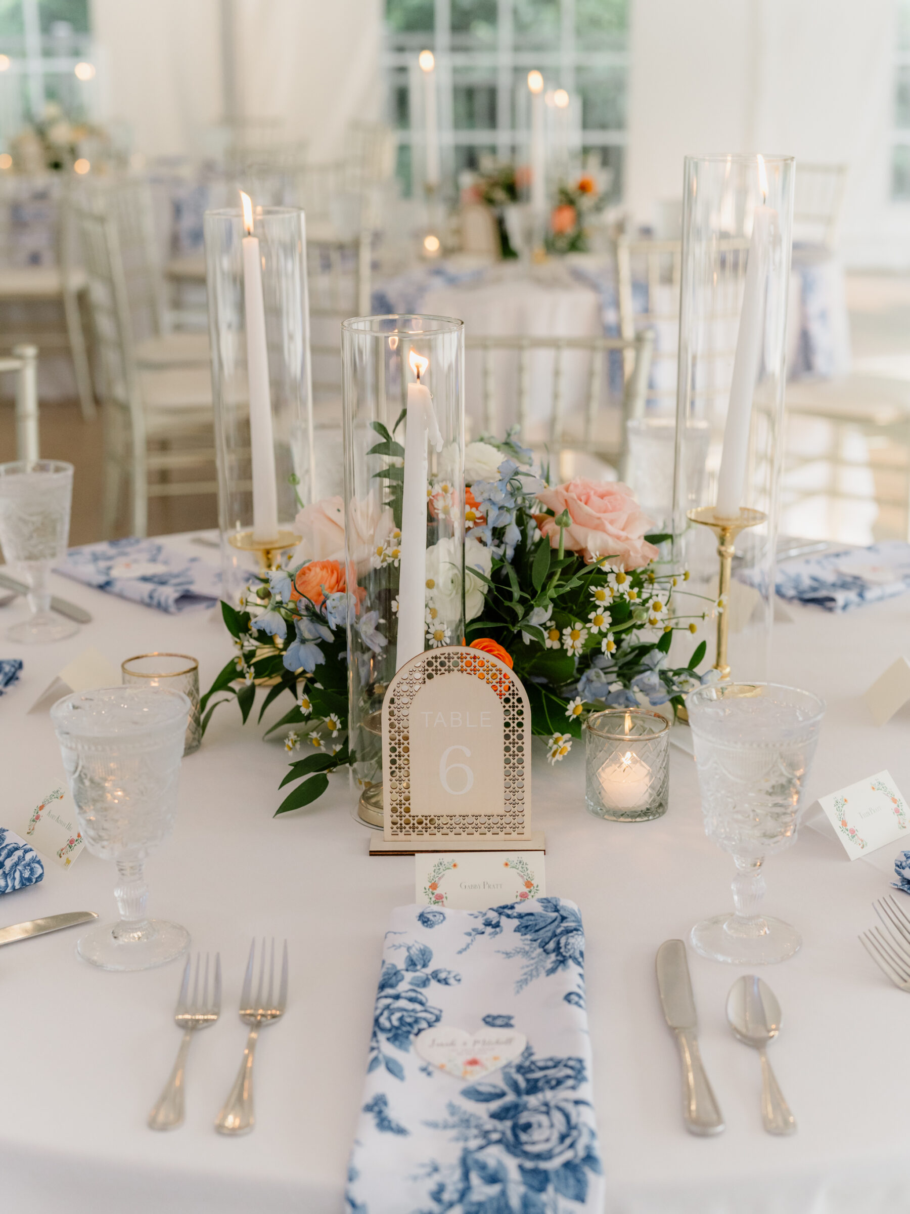 Elegant table setting with blue floral napkins, soft pastel centerpiece, and glowing taper candles surrounding a table six sign.