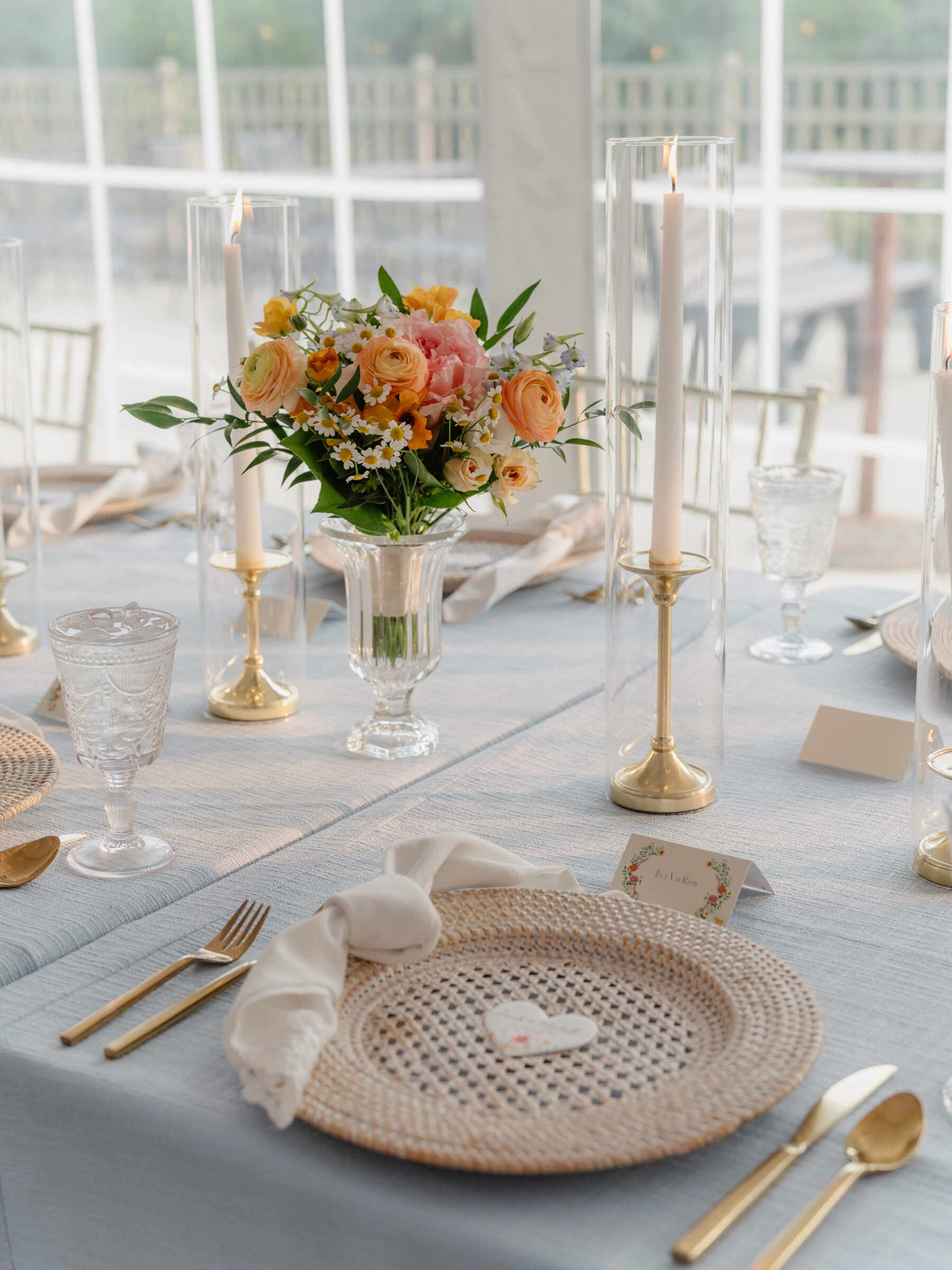 Elegant reception table with woven charger, blue linen, pastel blooms, and glowing taper candles.