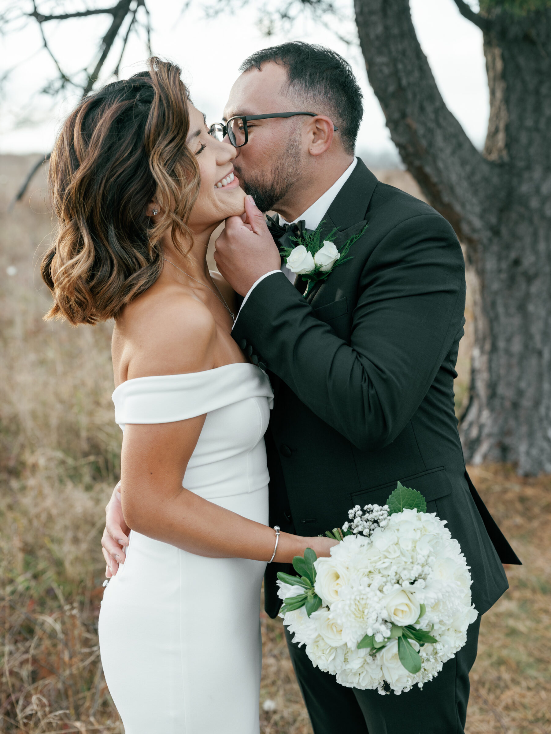 Groom gently kissing his bride’s cheek as she smiles, holding a white floral bouquet in an autumn meadow at Tanner’s Ridge.