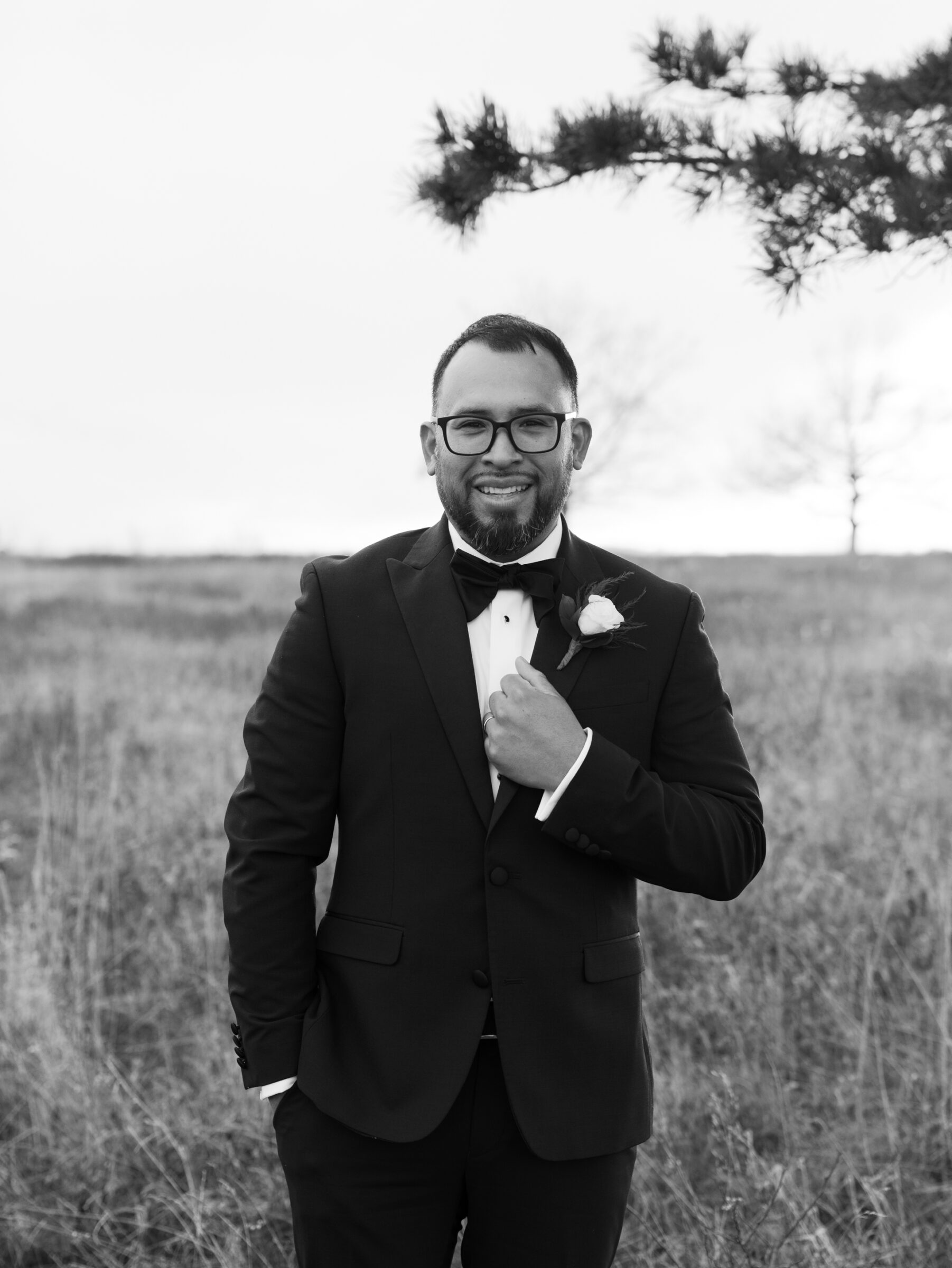 Black and white portrait of the groom smiling in a tuxedo, standing in an open meadow at Tanner’s Ridge.