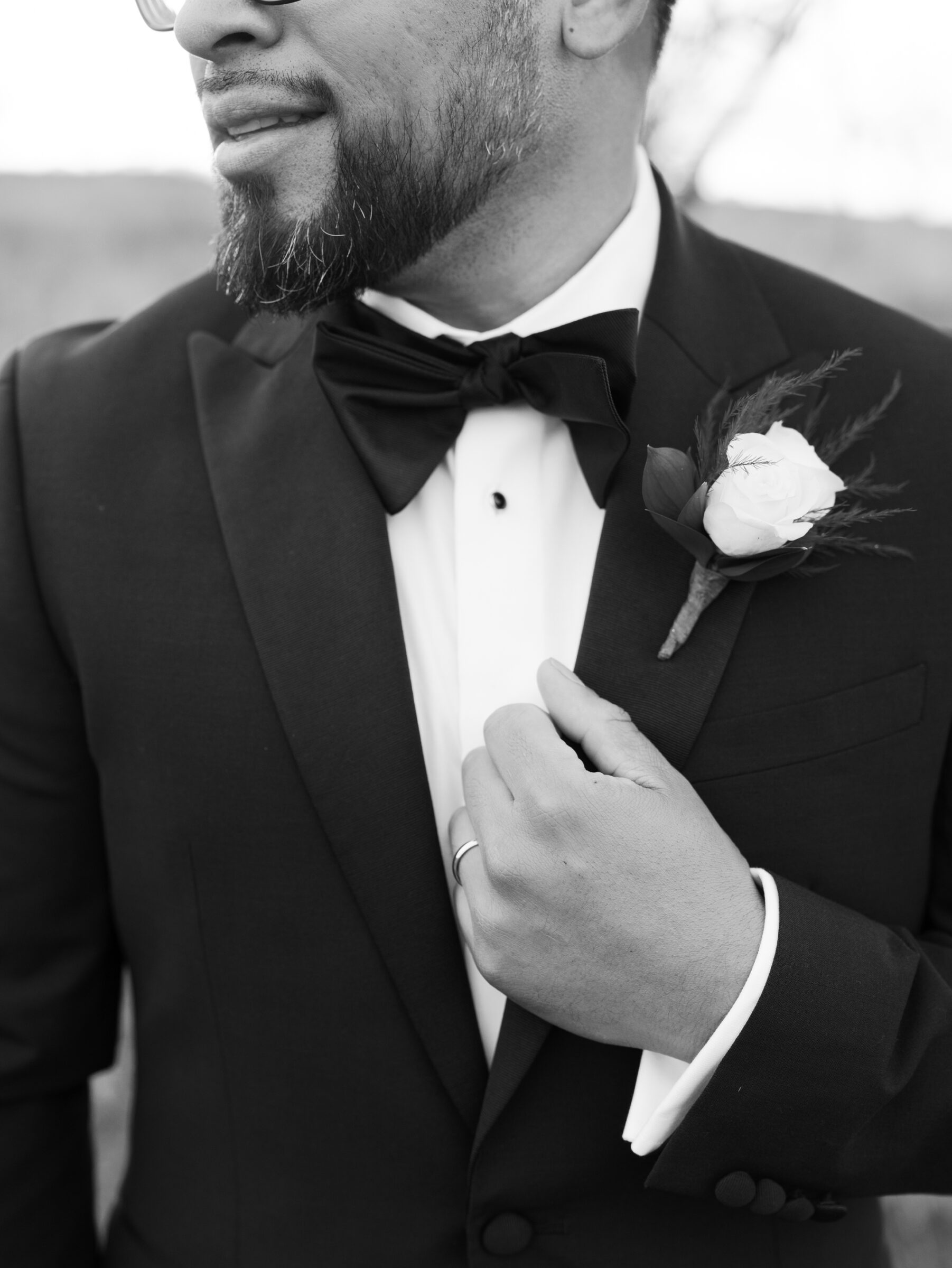 Black and white close-up of the groom adjusting his tuxedo jacket, showing his bow tie, boutonniere, and wedding band during mountain portraits.