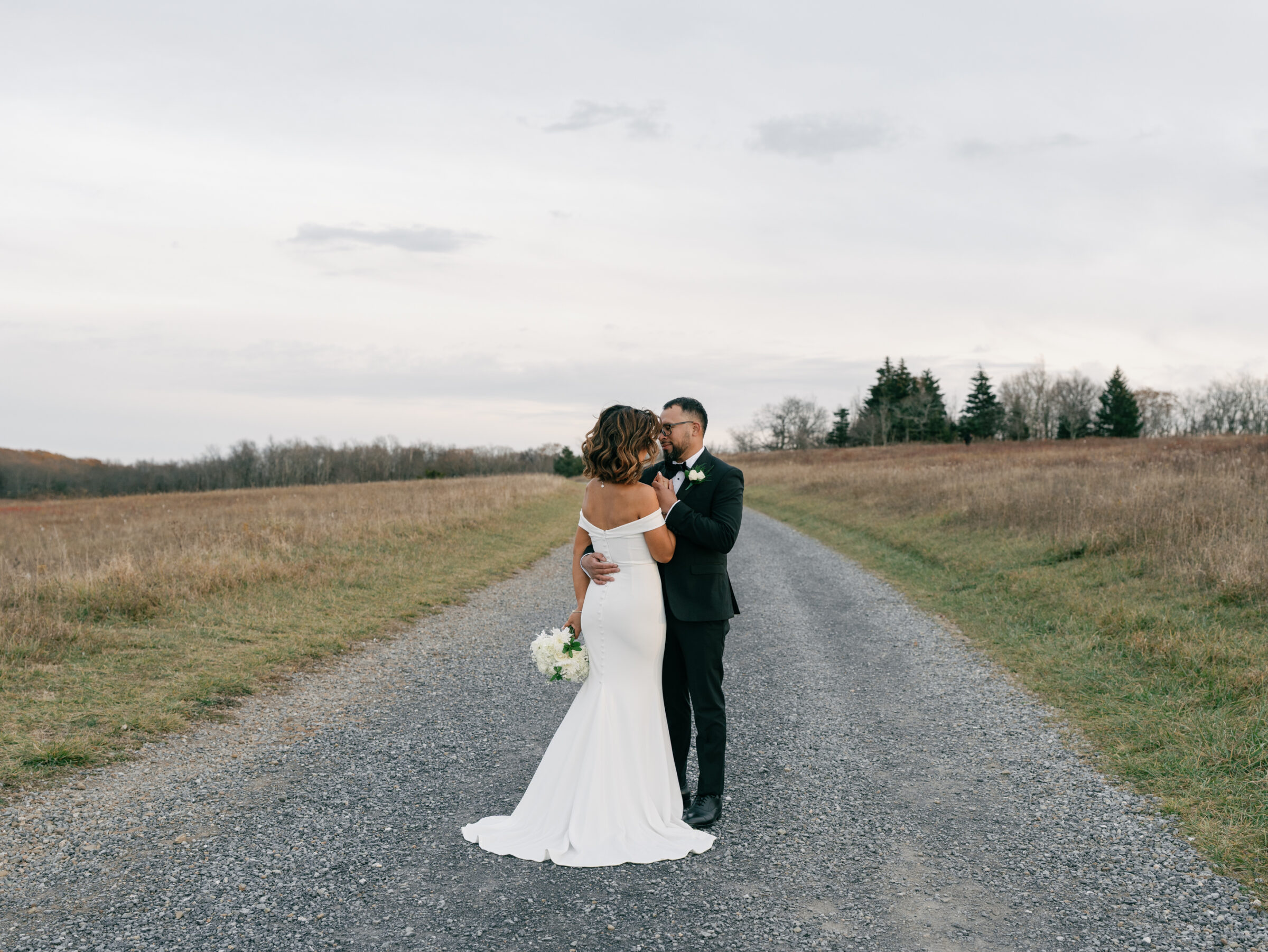 Bride and groom embracing on a gravel path surrounded by open meadows at Tanner’s Ridge during their autumn mountain portraits.