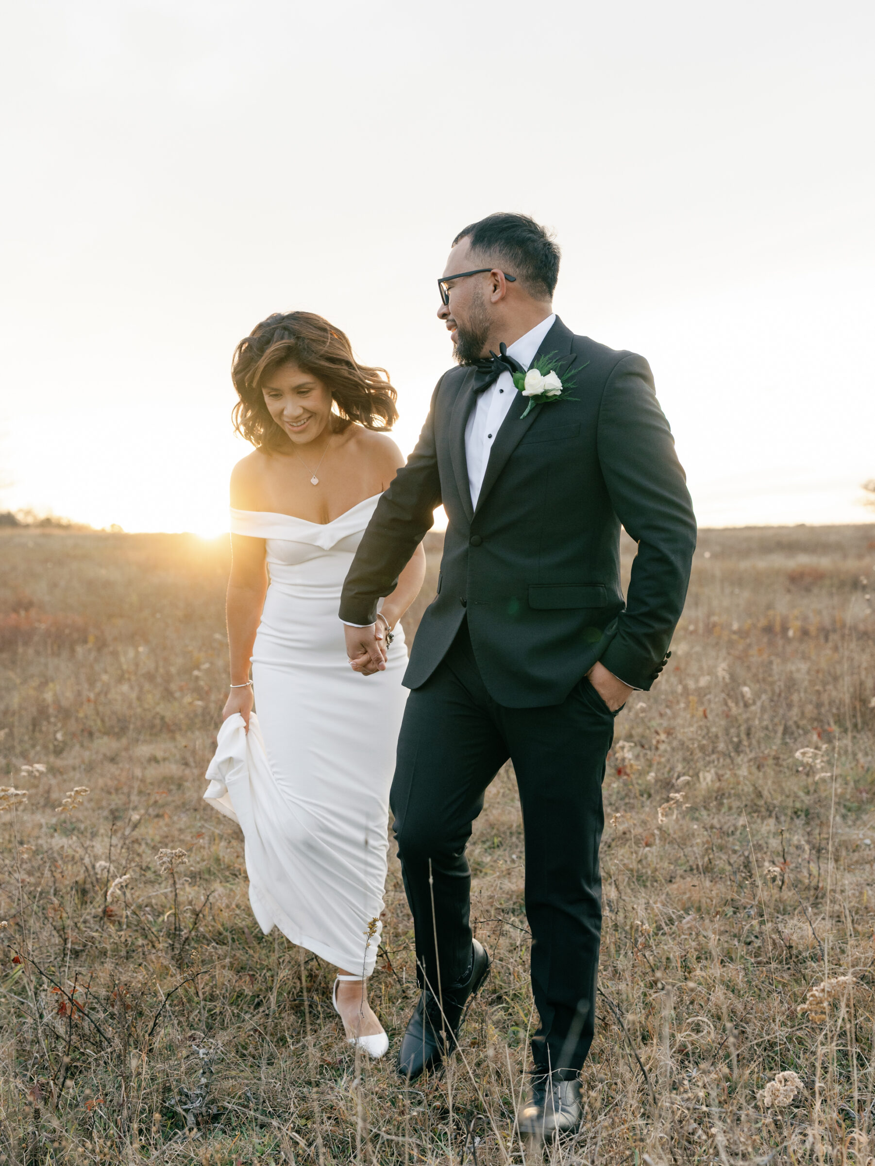 Skyline Drive Virginia Pictures | Bride and groom walking hand in hand through an autumn meadow at sunset, smiling as the light glows behind them at Tanner’s Ridge.